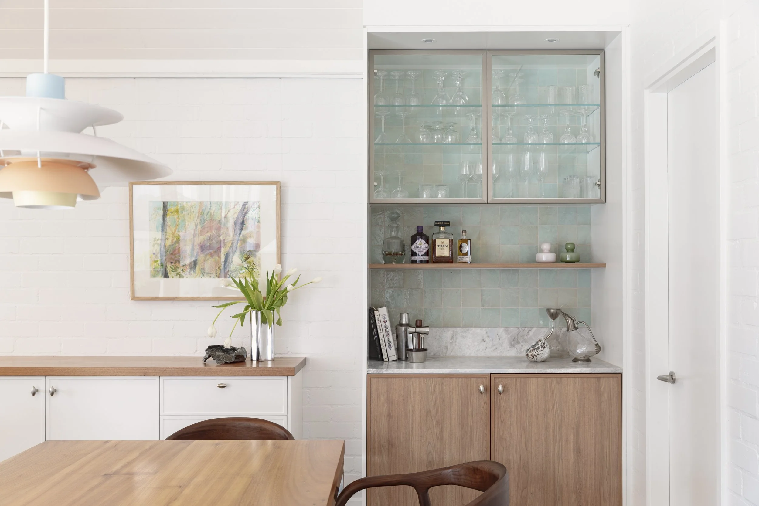 A corner of a modern kitchen with white brick walls, a wooden countertop, and a built-in glass-front cabinet with glassware. There is a painting on the wall, a vase with flowers, and decorative bottles on a shelf.