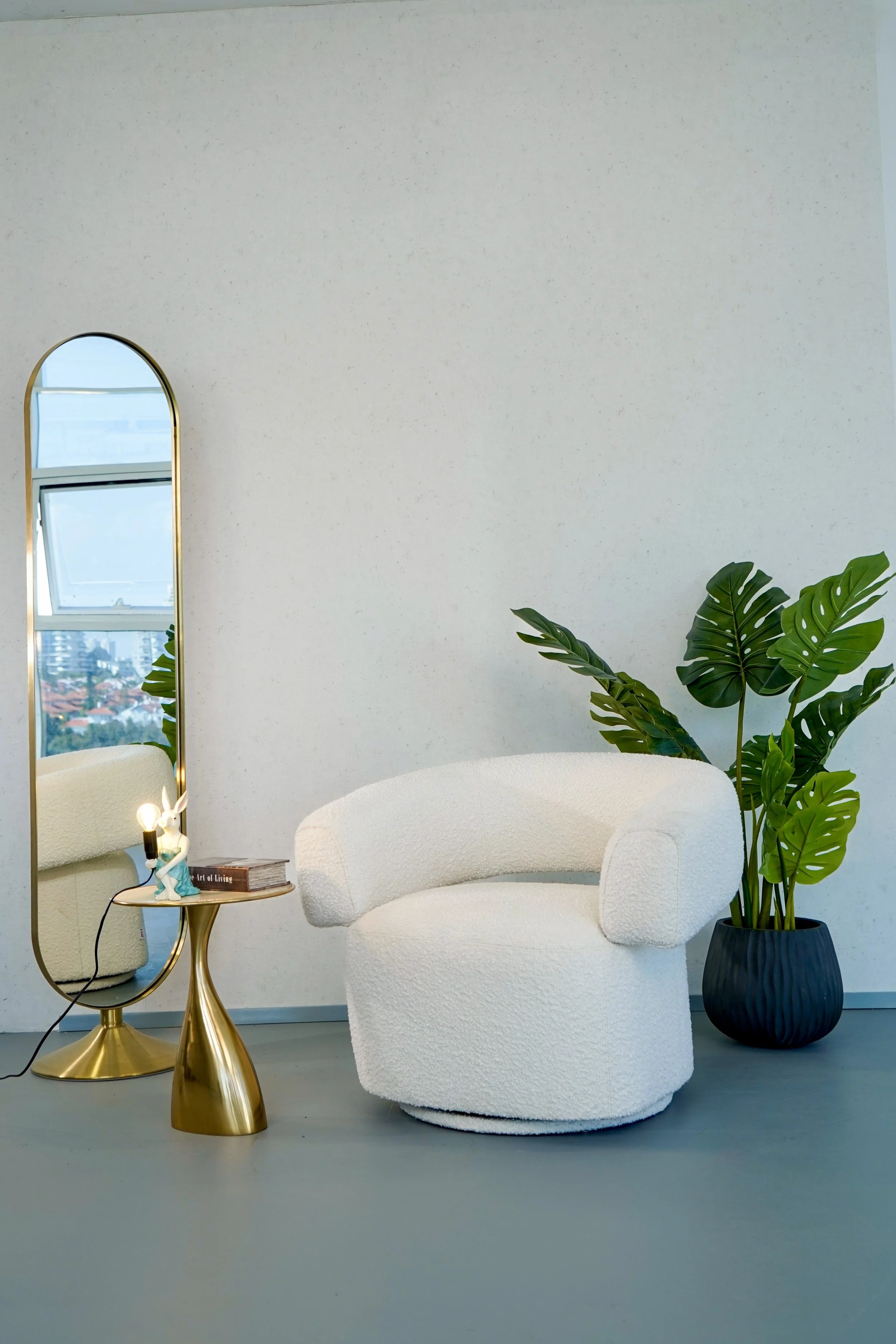 Minimalist interior with a white textured armchair, a tall black potted plant with large green leaves, a small round gold side table with a lamp and books, a tall mirror with a gold frame, and a window showing a city view.