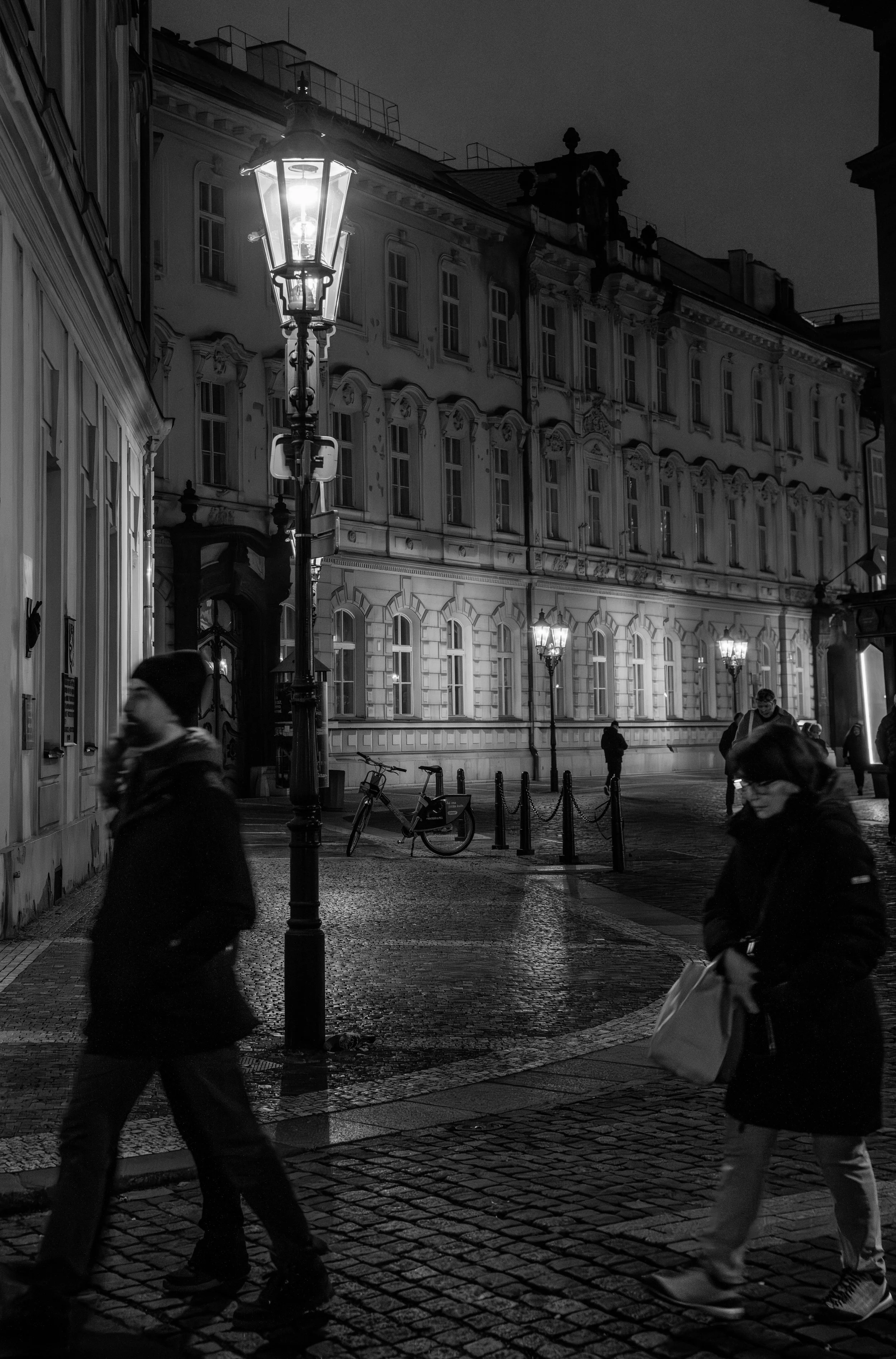 A black and white photo of a city street at night with pedestrians walking and streetlights illuminating the area.