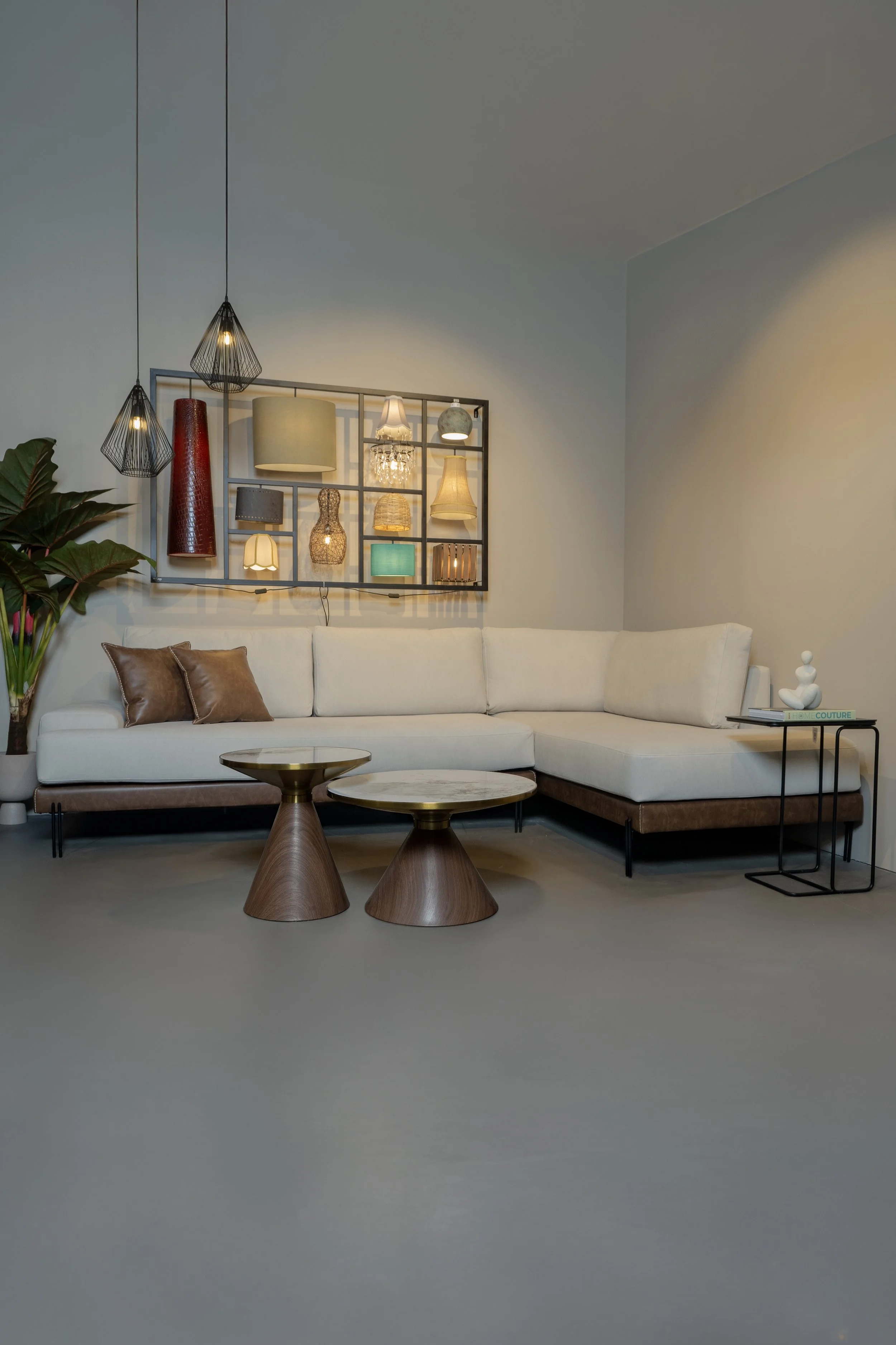 Modern living room with an off-white sectional sofa, two round wooden coffee tables, a black side table with decor and books, wall-mounted light fixtures, and a decorative wall art with various lamps and light fixtures.