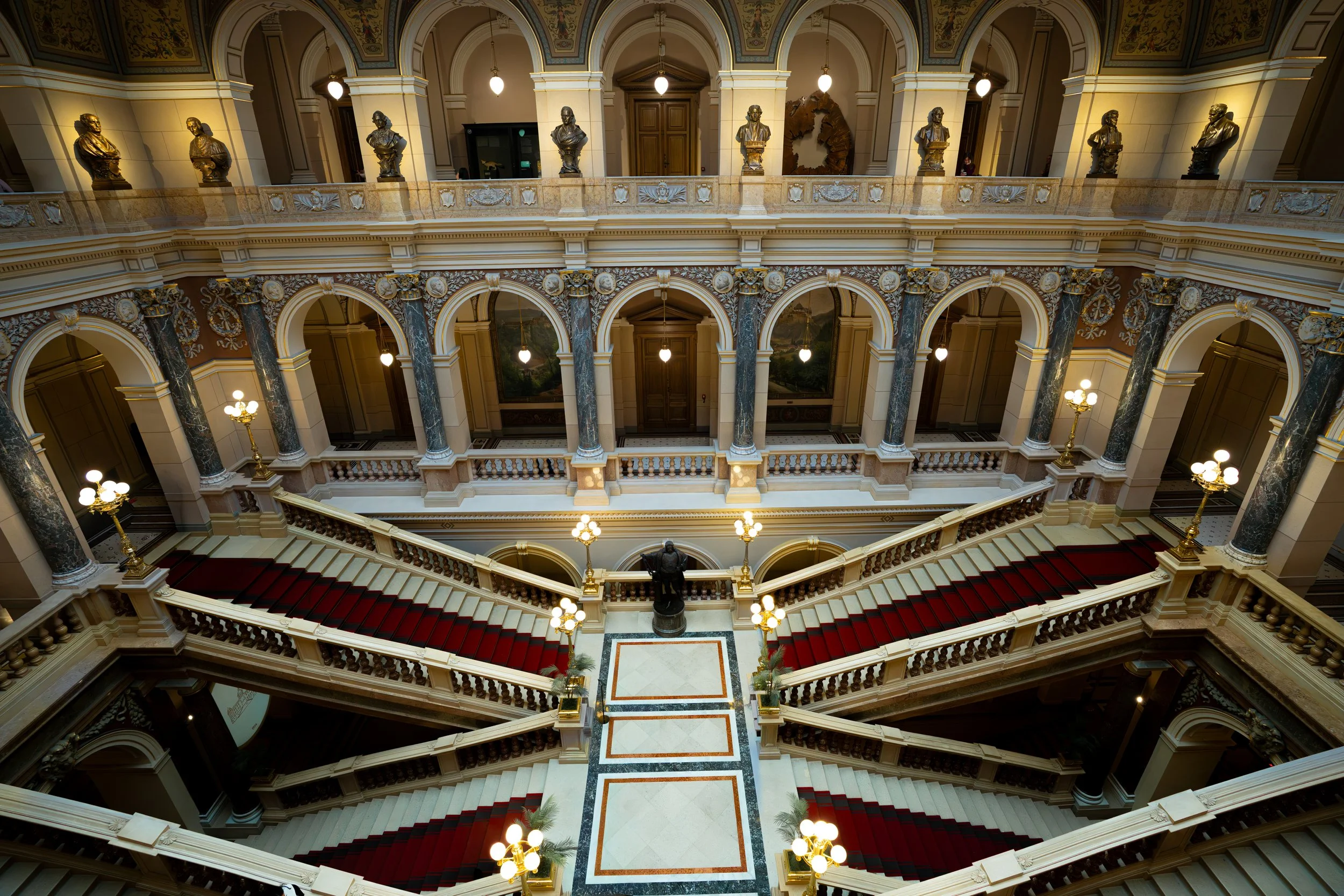 Elegant multi-story staircase with red-carpeted steps, ornate columns, golden chandeliers, and bust sculptures along the upper balcony in a grand interior space.