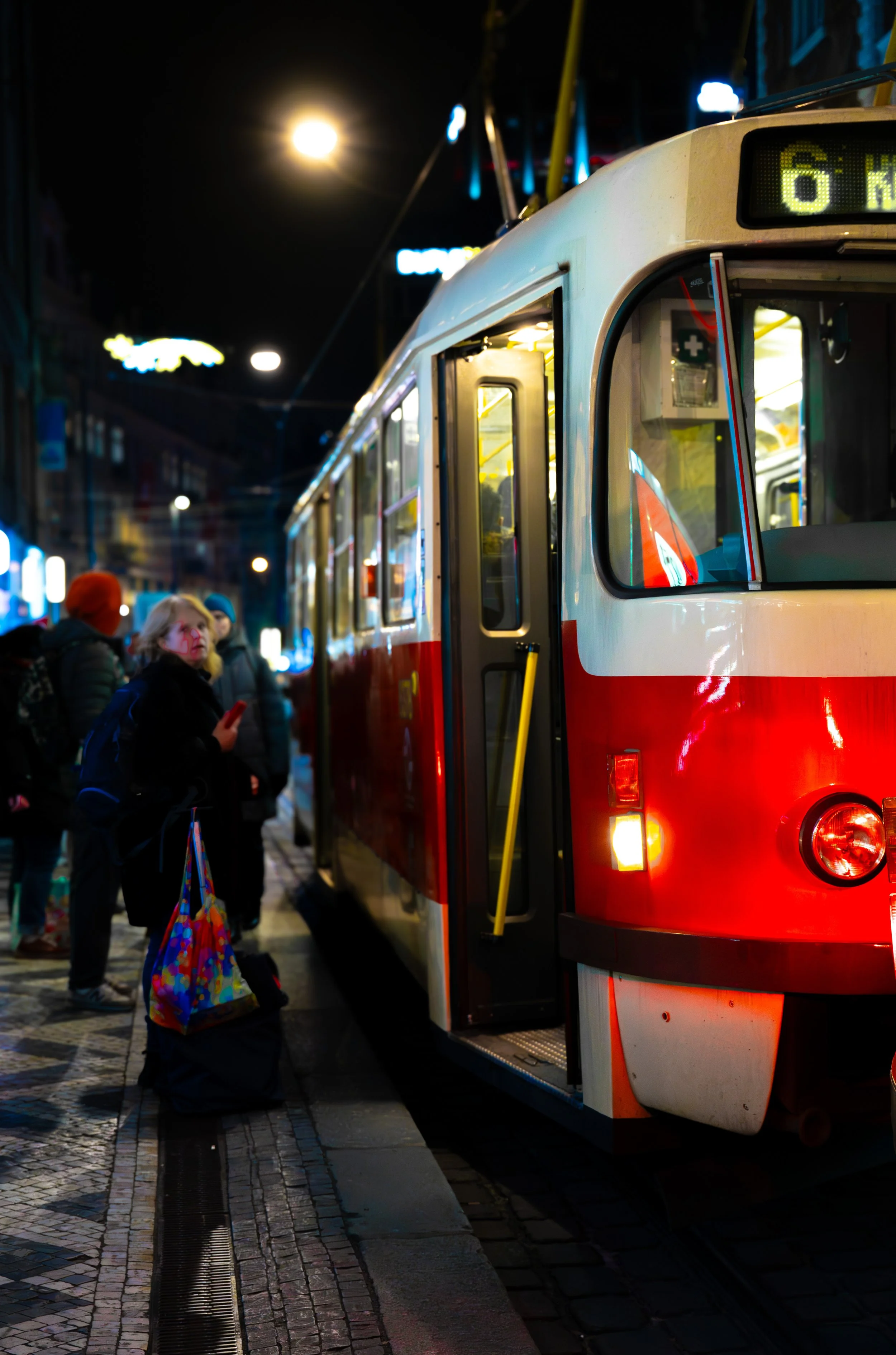 Nighttime scene of a red and white tram with passengers waiting at a city stop, illuminated by streetlights and surrounding buildings.