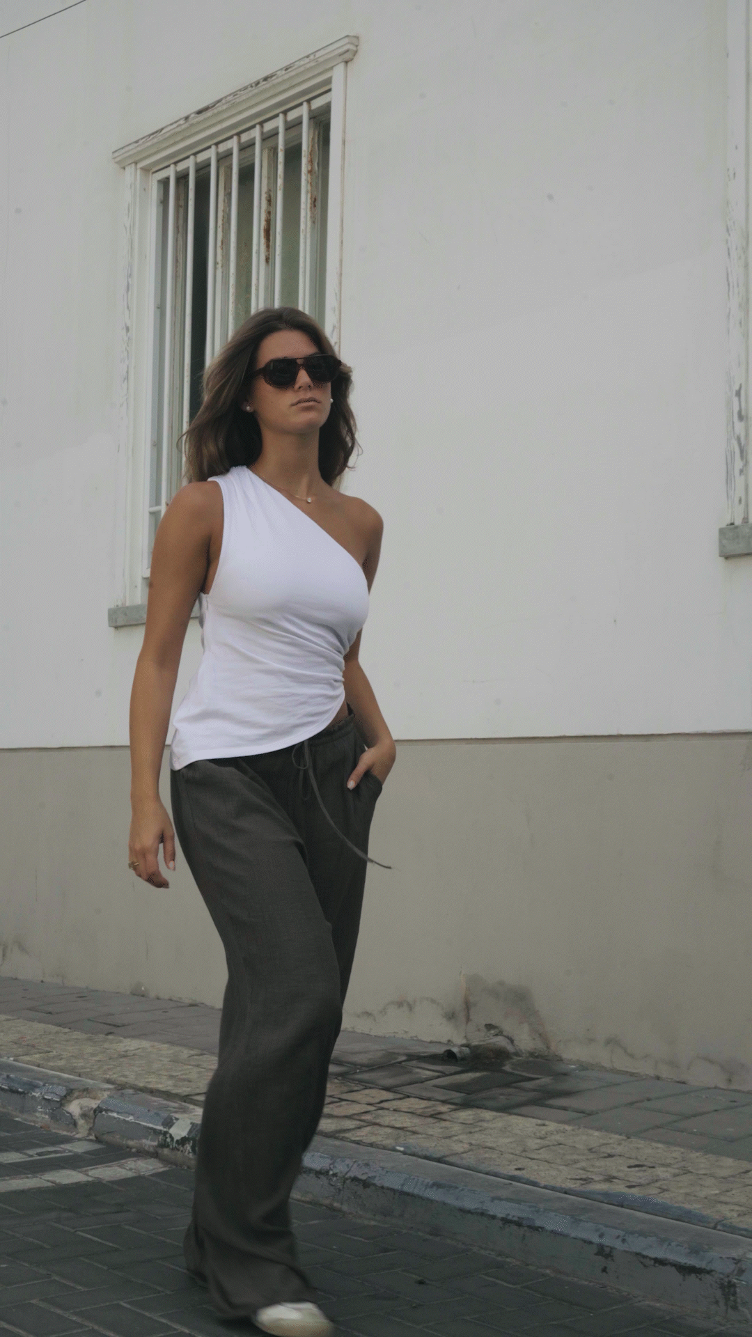 A young woman with dark hair, wearing sunglasses, a white one-shoulder top, gray pants, and white shoes, walking along a sidewalk next to a white building with a barred window.