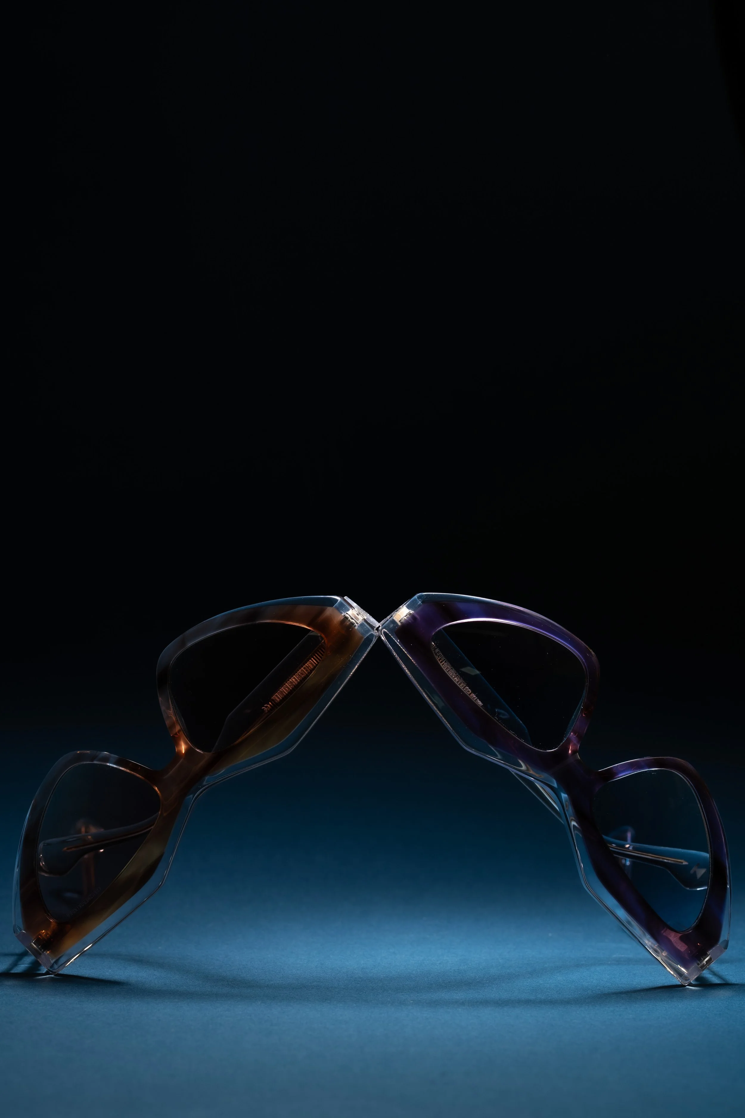 Two pairs of eyeglasses with colorful frames balancing with their temples touching, on a blue surface against a dark background.