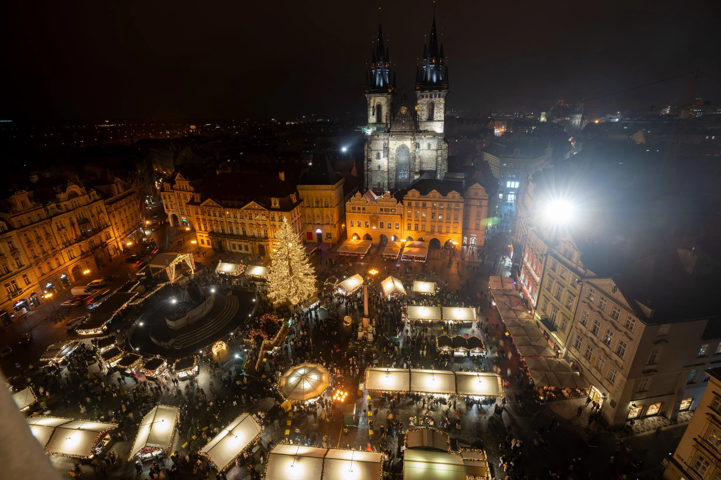 Nighttime scene of a Christmas market in a historic European city square, with a large decorated Christmas tree, market stalls, and a prominent Gothic church in the background.