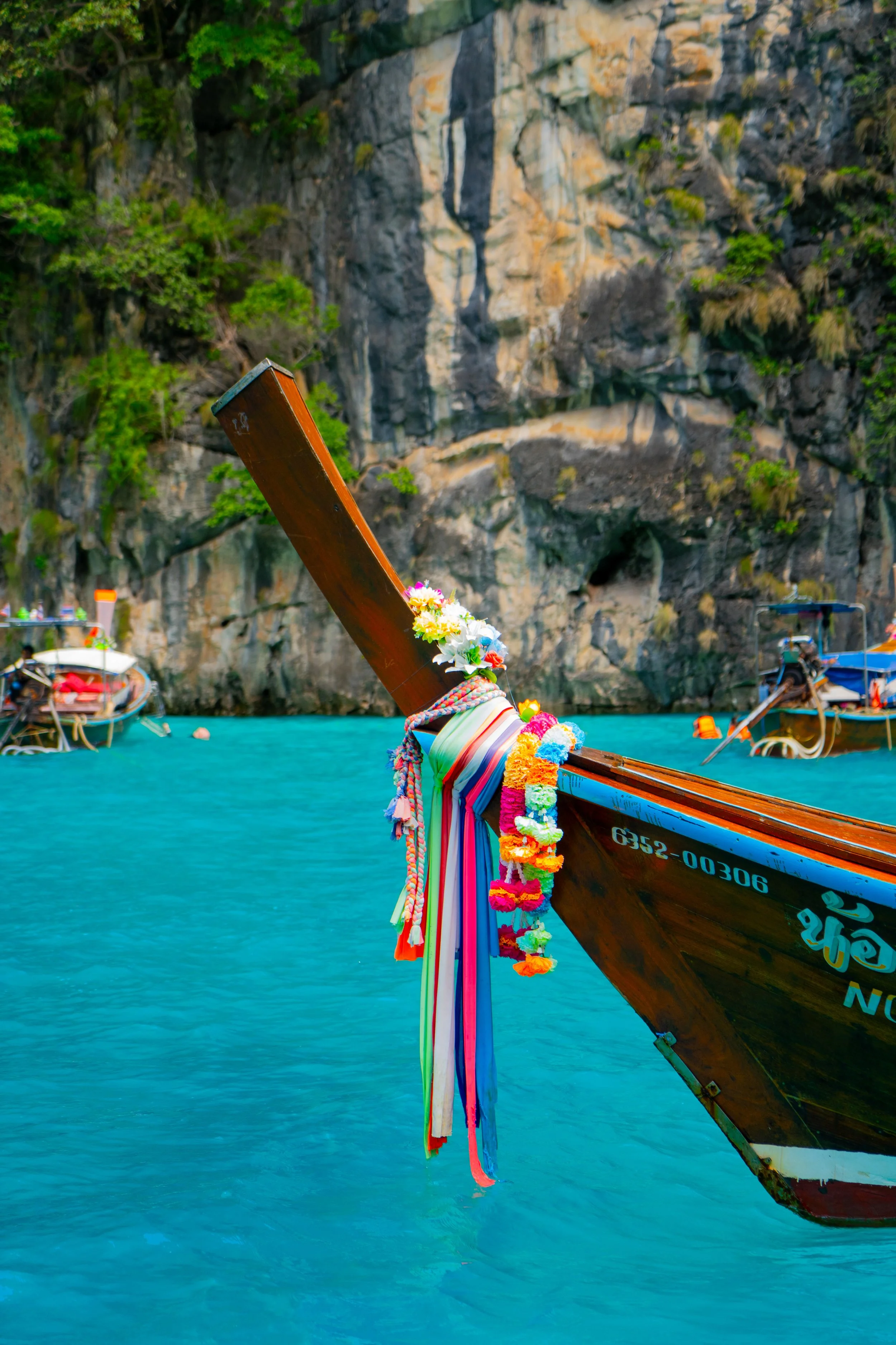 Traditional Thai longtail boat decorated with colorful fabric and flower garlands floating on turquoise water with limestone cliffs and other boats in the background.