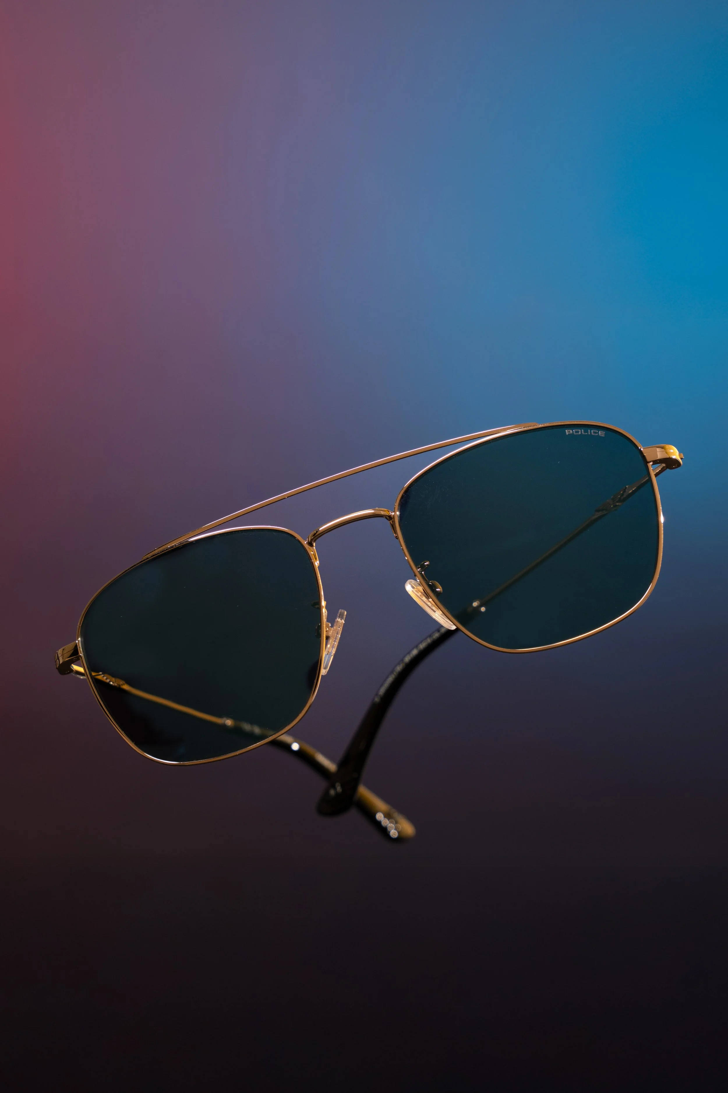 A pair of sunglasses with dark lenses and a thin gold frame against a gradient background of purple and blue.