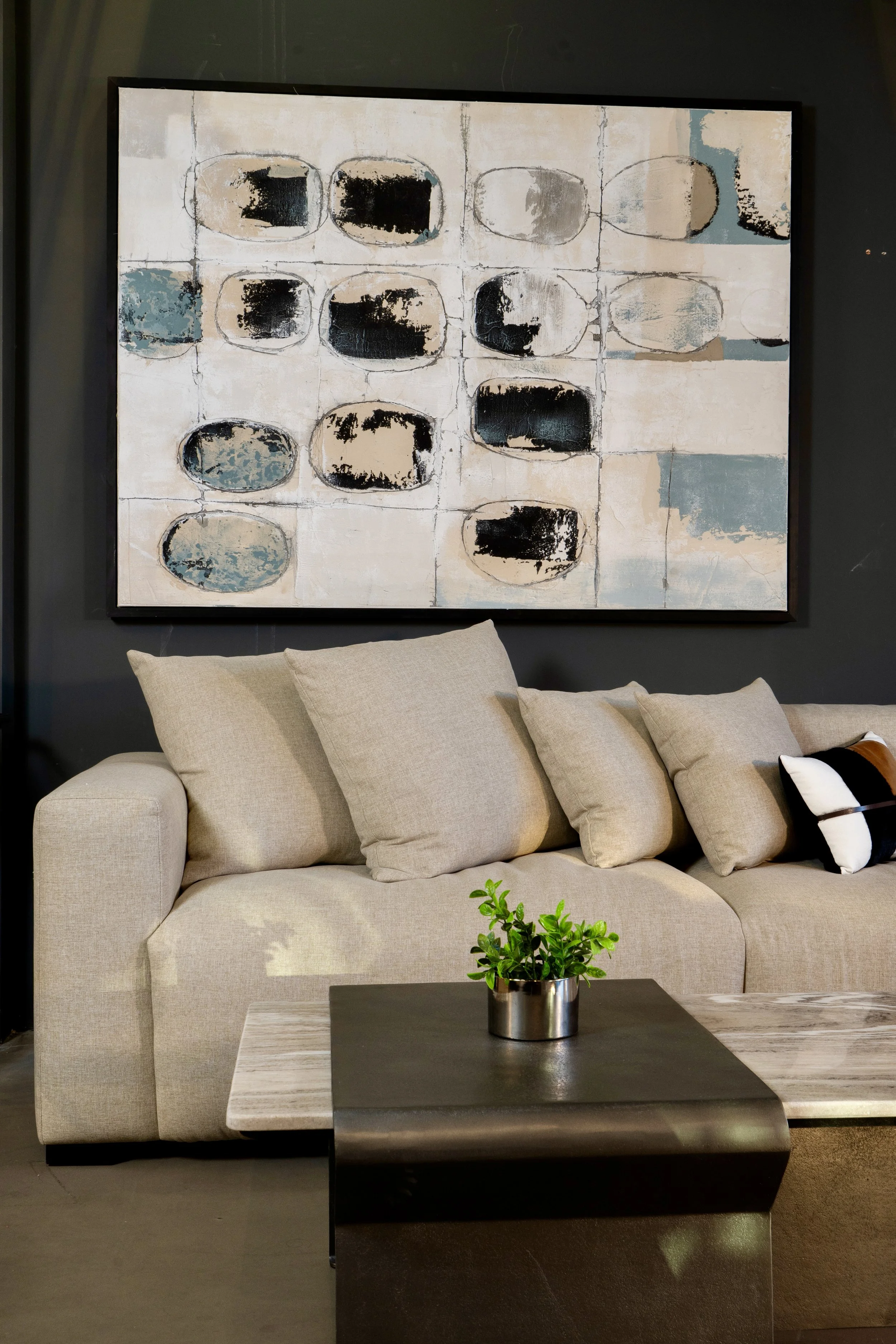 A beige sofa with multiple cushions placed against a dark-colored wall, with a large modern abstract painting hanging above it. In front of the sofa, there is a dark-colored coffee table with a small green potted plant.