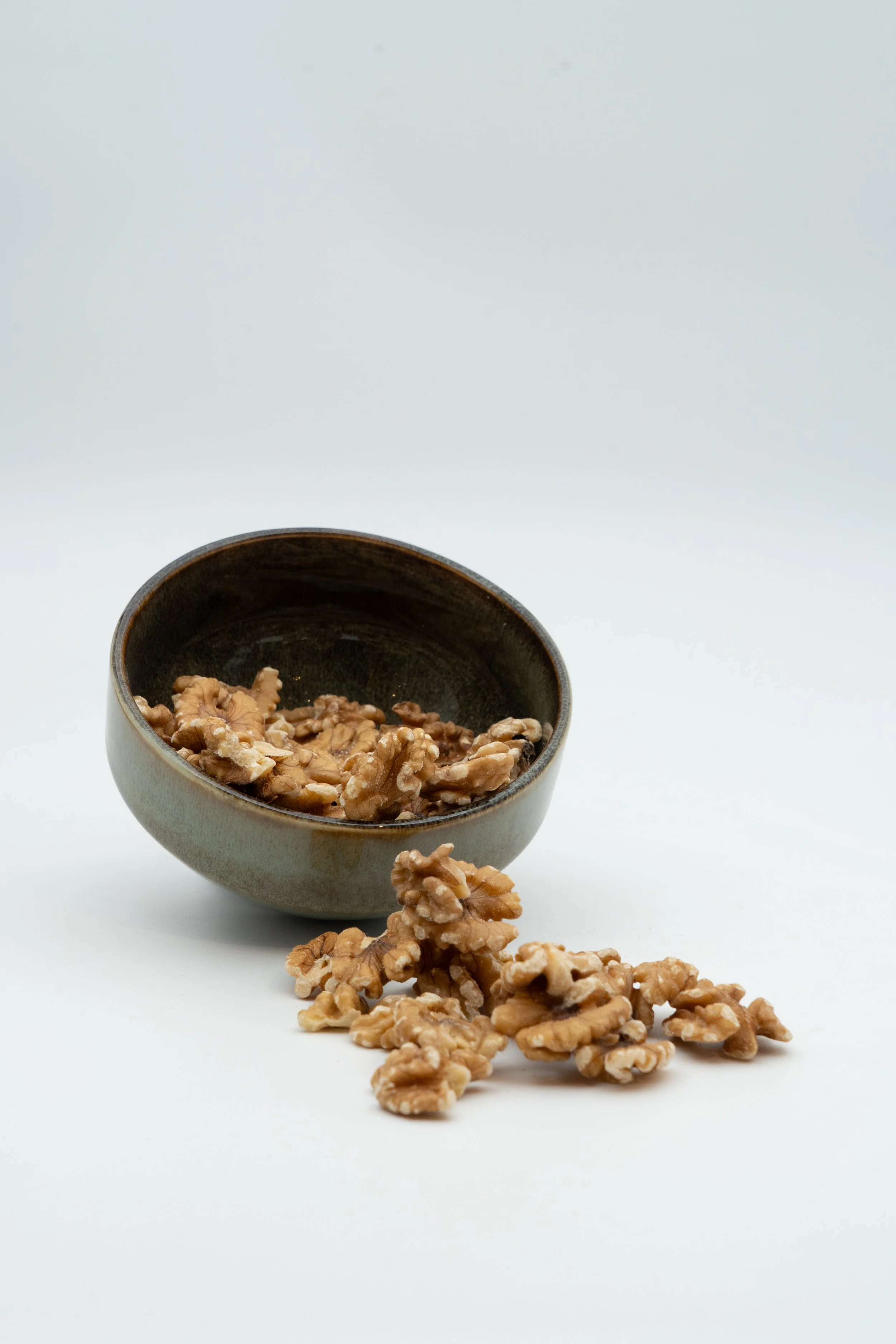 A small brown bowl filled with walnuts, with some walnuts spilled out in front, against a plain white background.