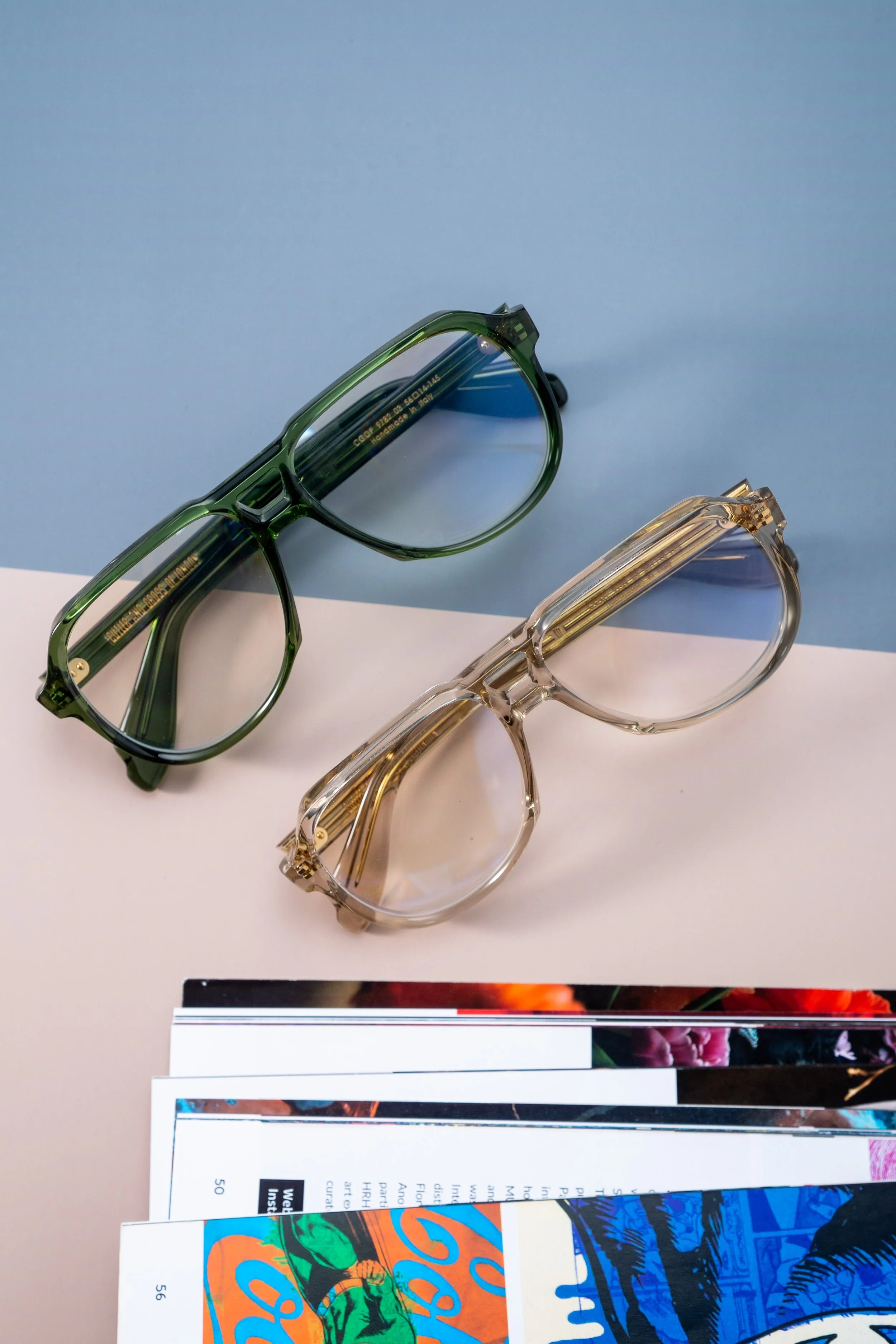 Two pairs of sunglasses, one green and one clear, resting on a table with magazines underneath. The background is split between blue and white surfaces.