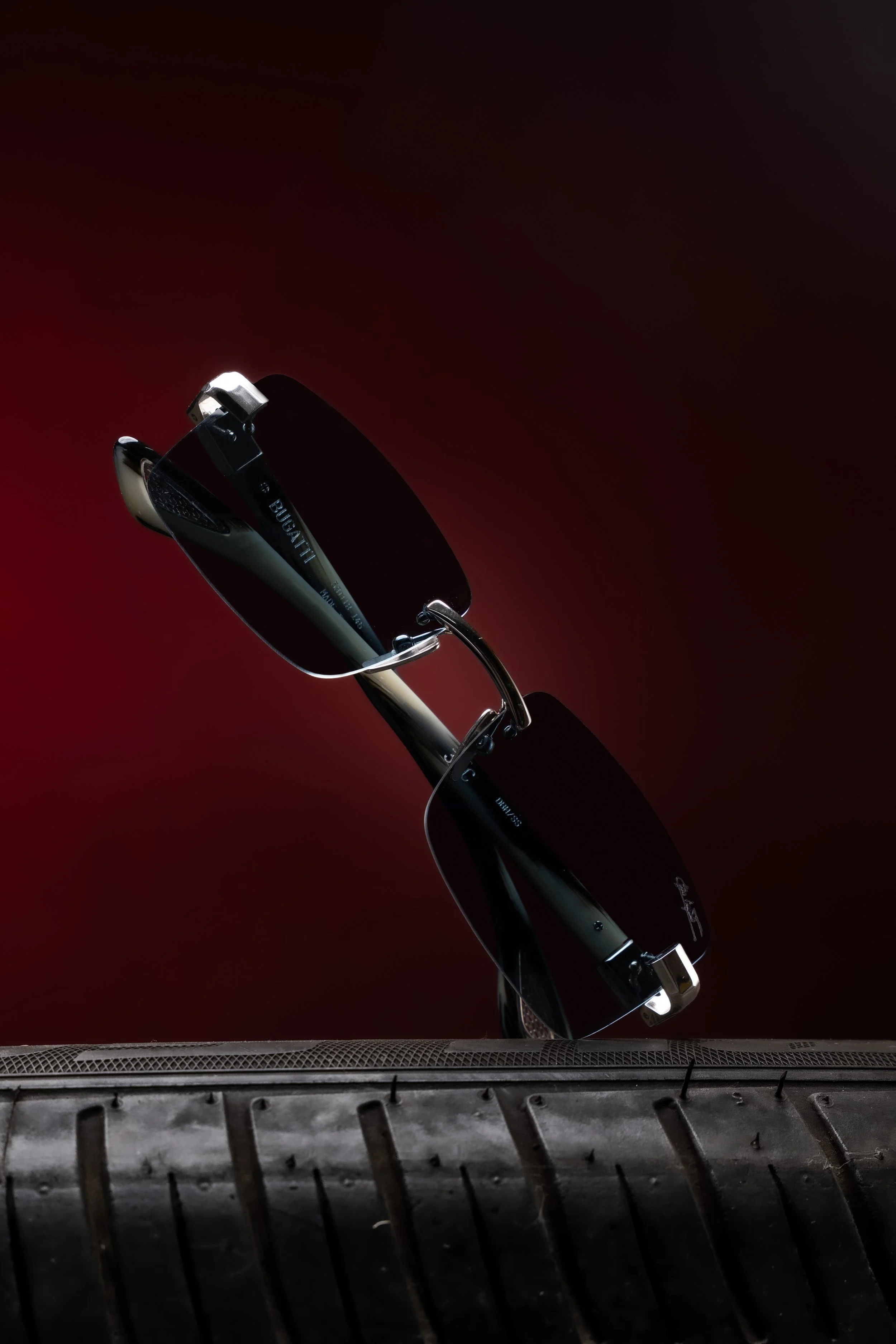 A pair of black sunglasses with the brand logo on the temple, positioned on a tire with a red background.