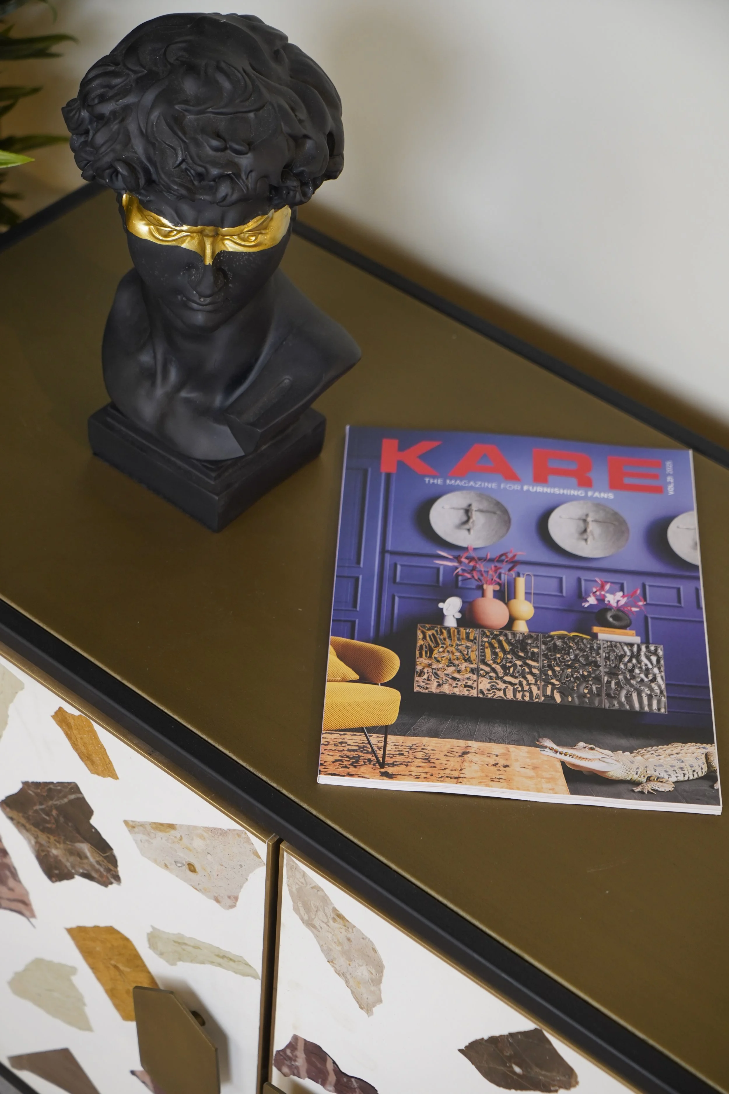 A black bust sculpture with curly hair and a gold blindfold on a desk next to a magazine titled 'KARE' featuring interior decor, with a crocodile figurine partially visible at the edge of the desk.