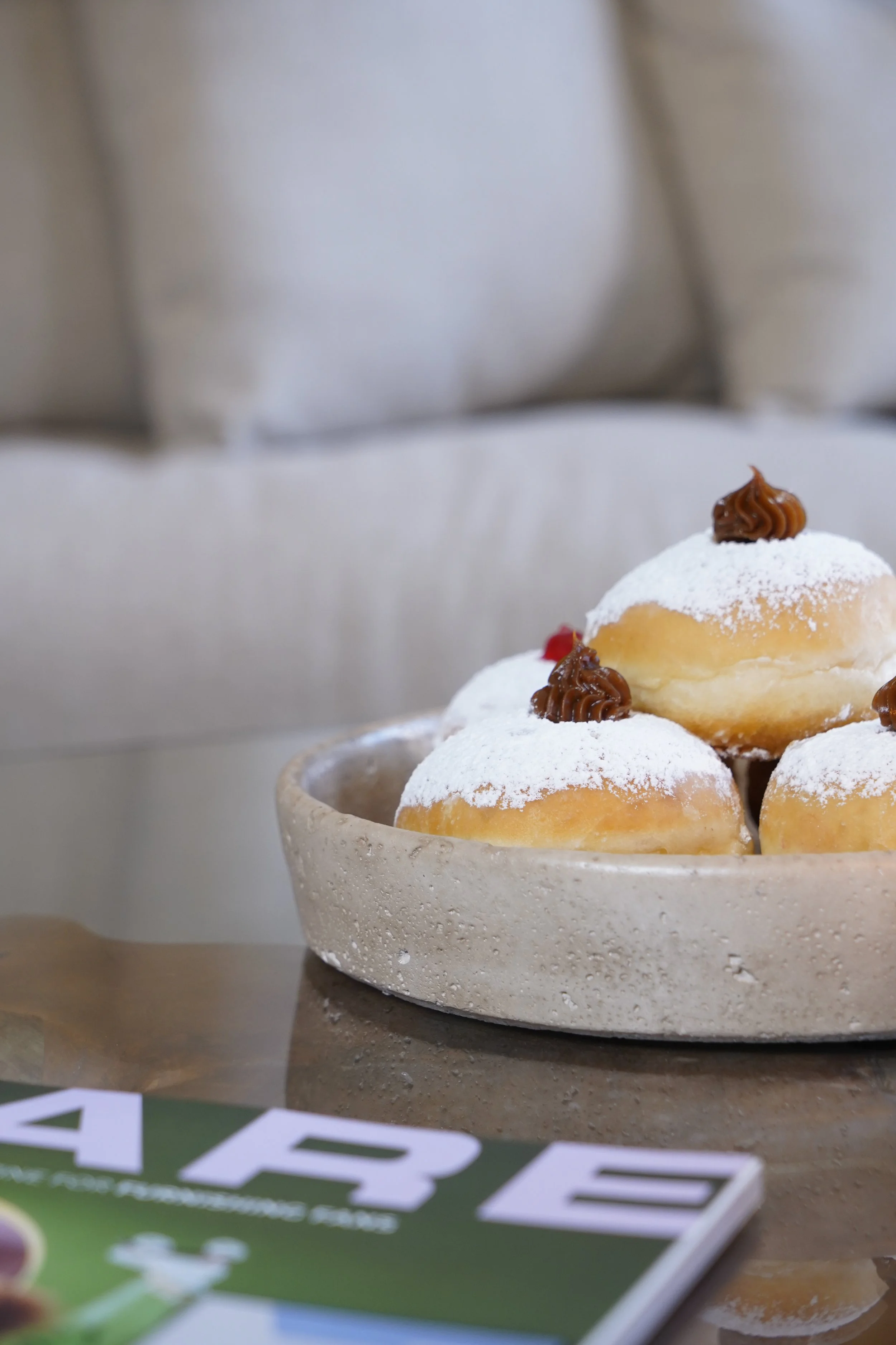 A beige ceramic bowl filled with powdered sugar-covered mini donuts topped with chocolate frosting on a wooden table.