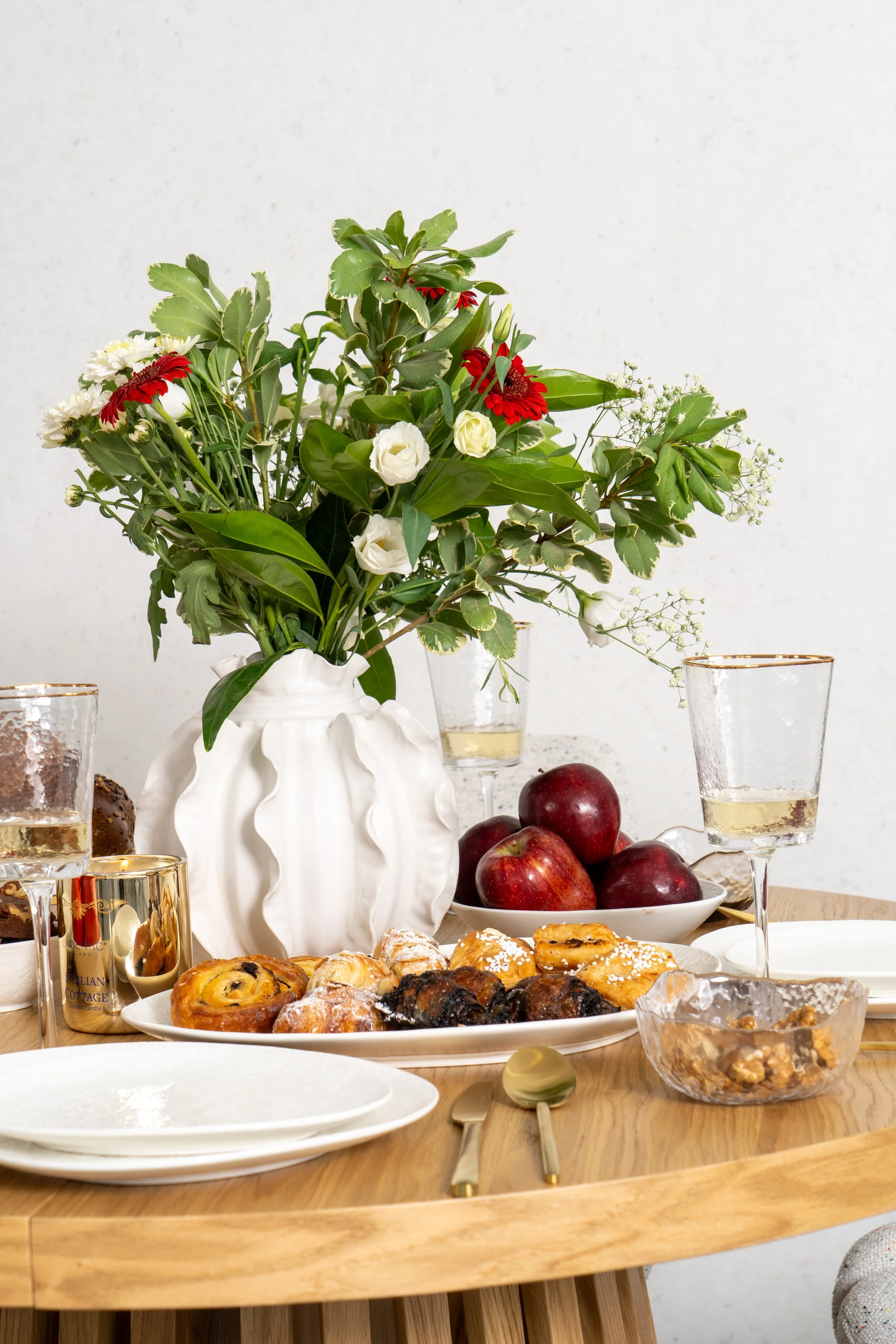 A table set with a vase of mixed flowers, glasses of white wine, apples, pastries, and a bowl of walnuts.