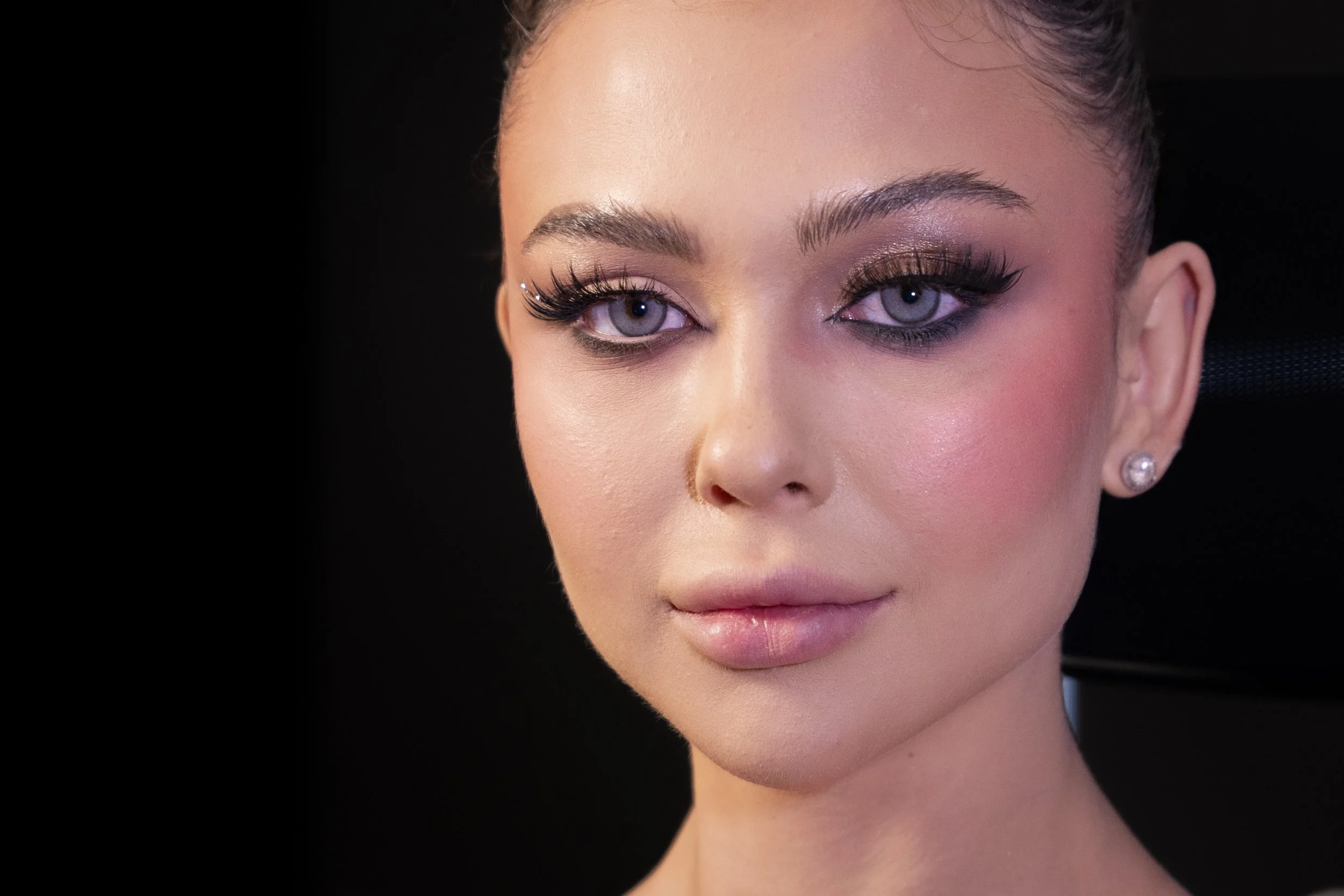 Close-up of a woman with fair skin, styled makeup, long eyelashes, and piercing blue eyes. She is wearing diamond stud earrings and has light pink lipstick and shimmery eye shadow.