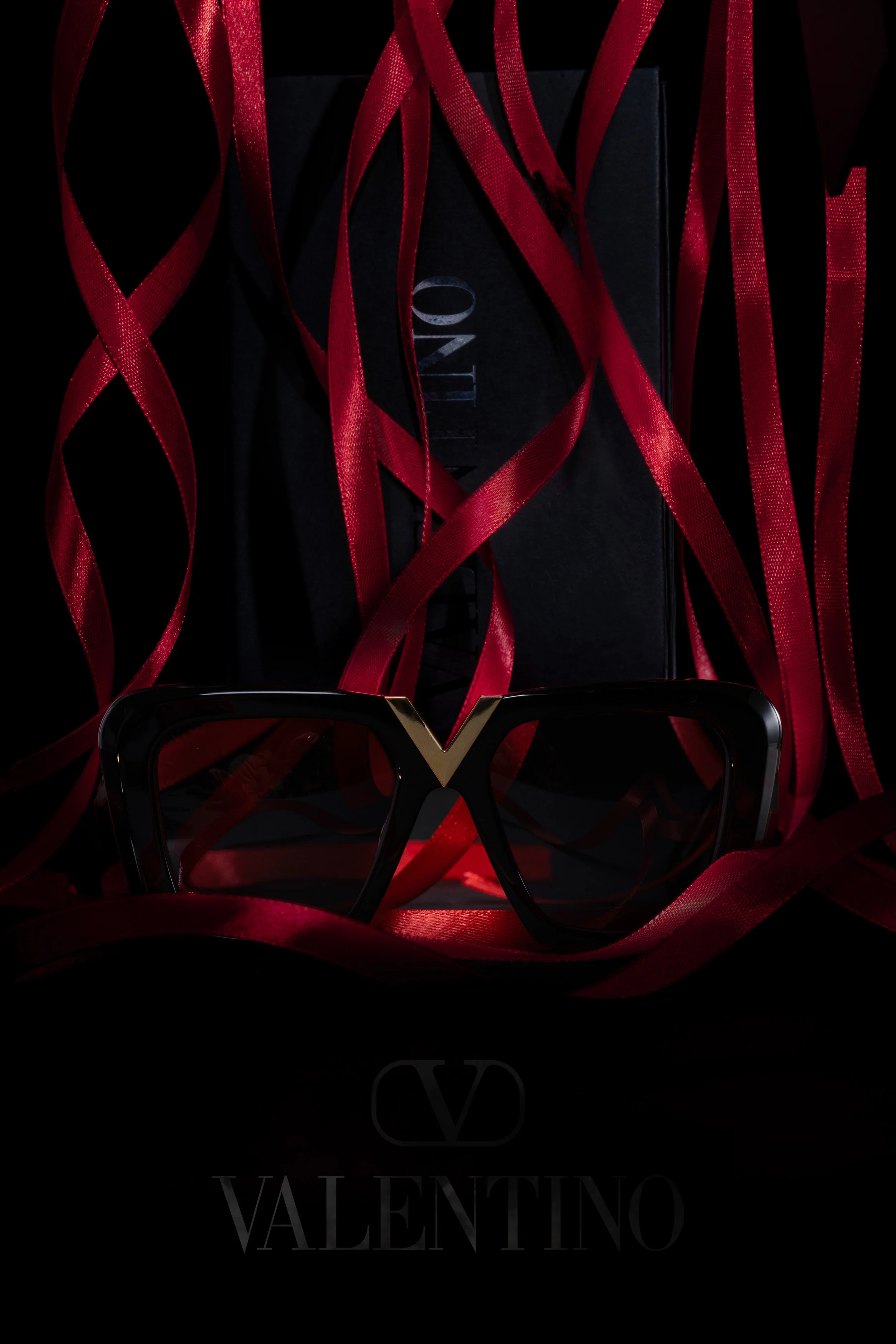 A pair of black Valentino sunglasses with a red and black Valentino box in the background, surrounded by red ribbons.