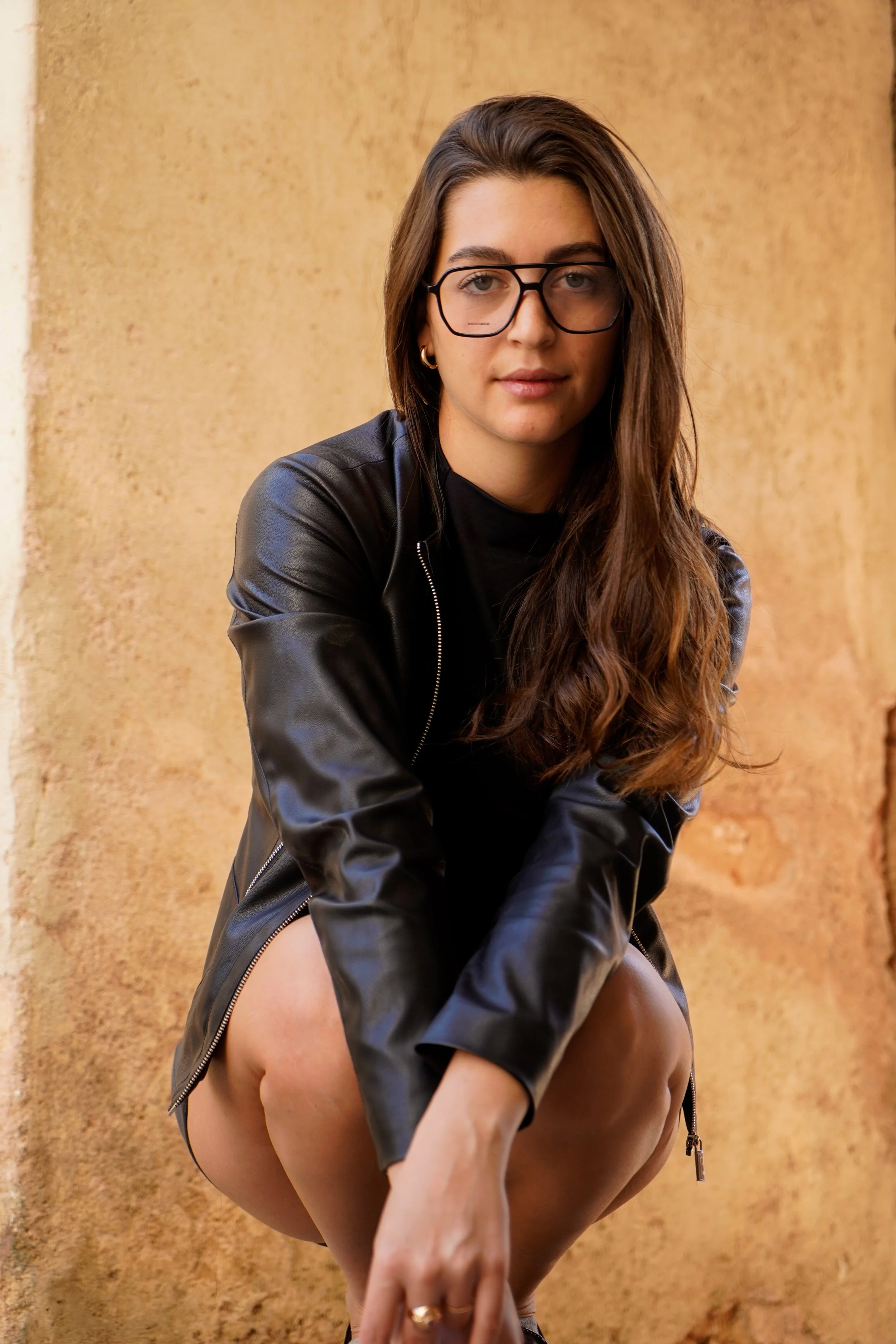 Young woman with long brown hair, wearing glasses, a black leather jacket, and black shirt, squatting outdoors against a textured beige wall.