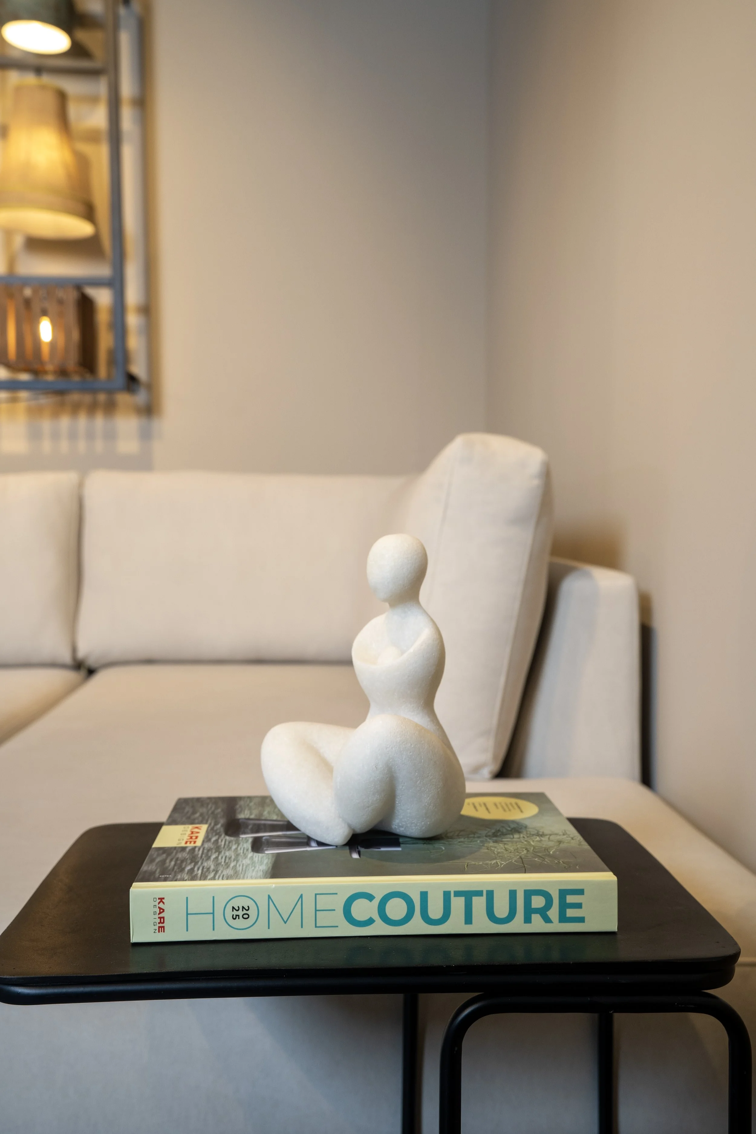 A white abstract sculpture of a seated woman on top of a home design magazine titled 'Home Couture,' placed on a black table in a room with beige sofas and a wall mirror.