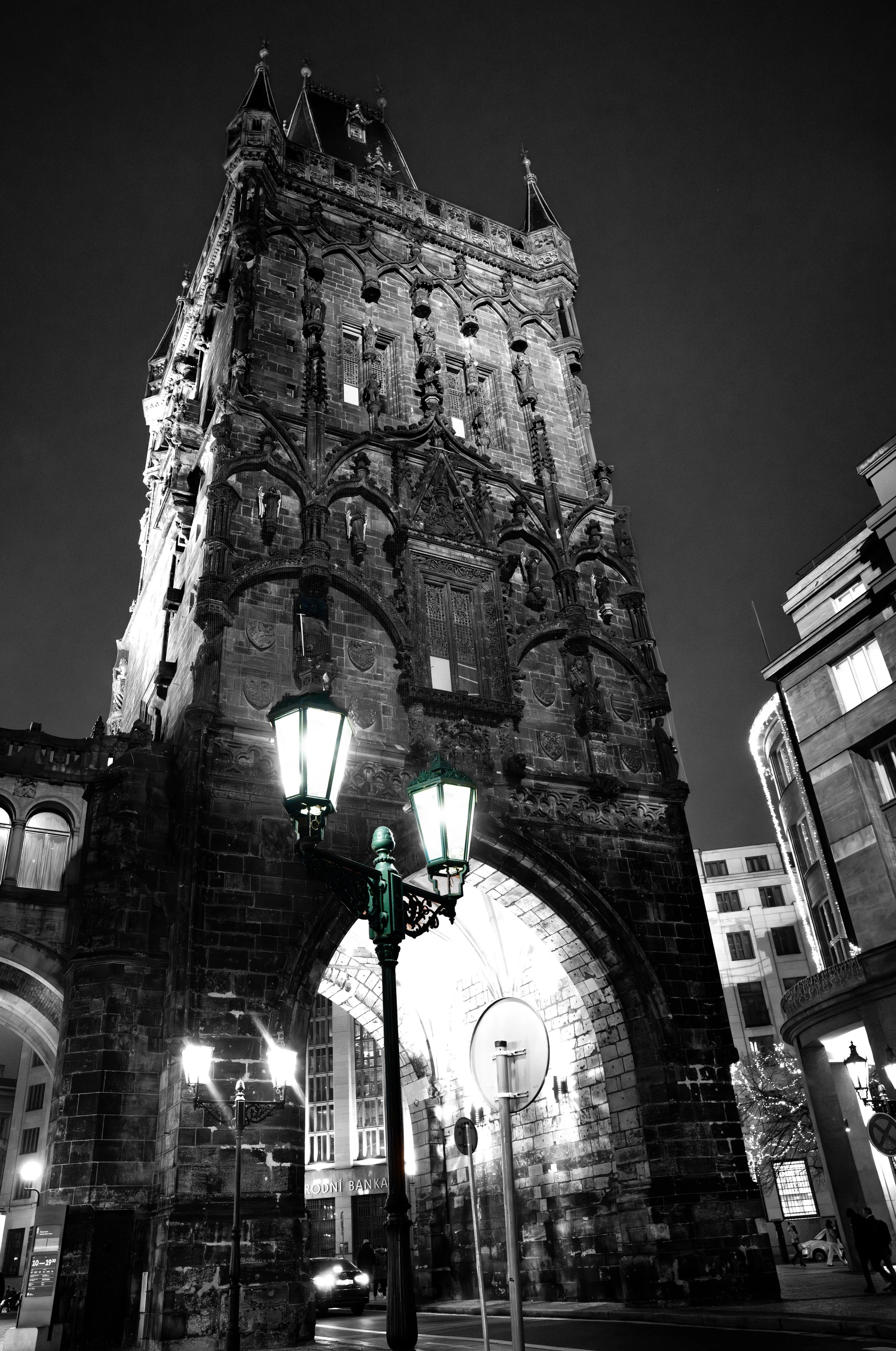 Night view of a historic clock tower with Gothic architecture, illuminated street lamps, and surrounding modern buildings in an urban setting.