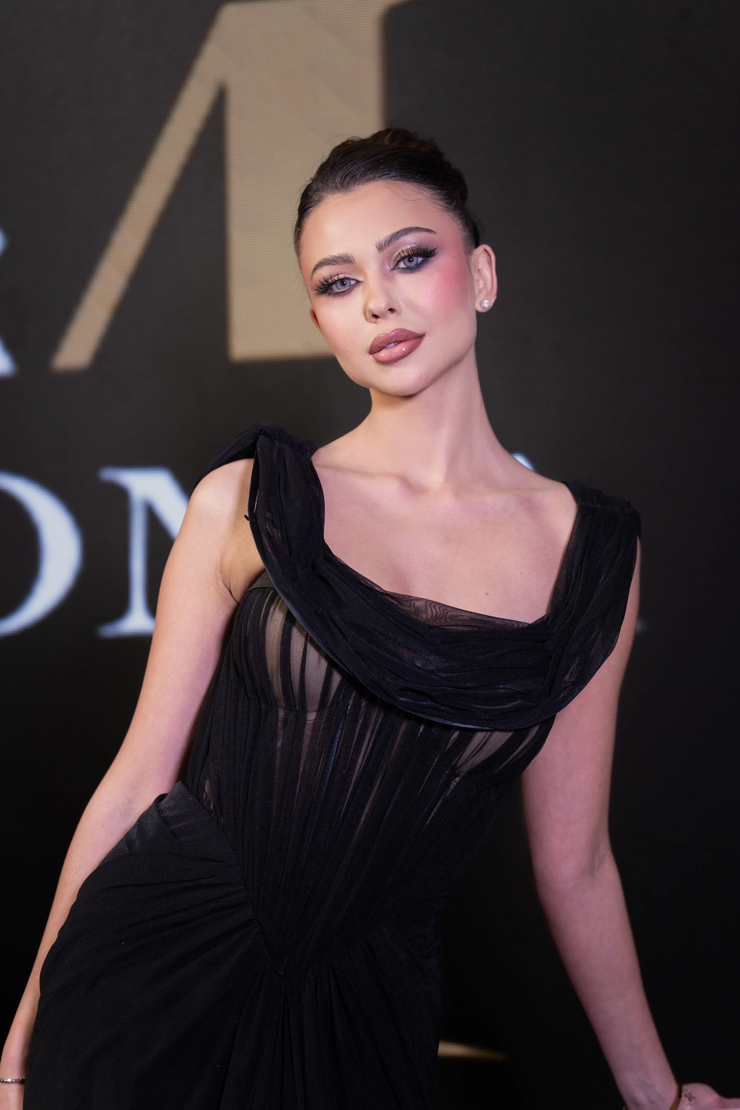 A woman with dark hair styled in an updo posing in a black dress with sheer and ruched details at a formal event, with a dark background and partially visible text.