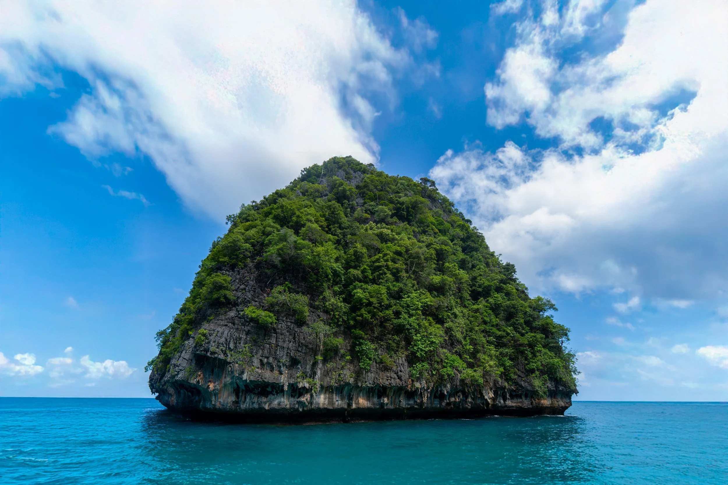 A large, lush, green island with steep rocky sides rising out of calm blue ocean water under a partly cloudy sky.