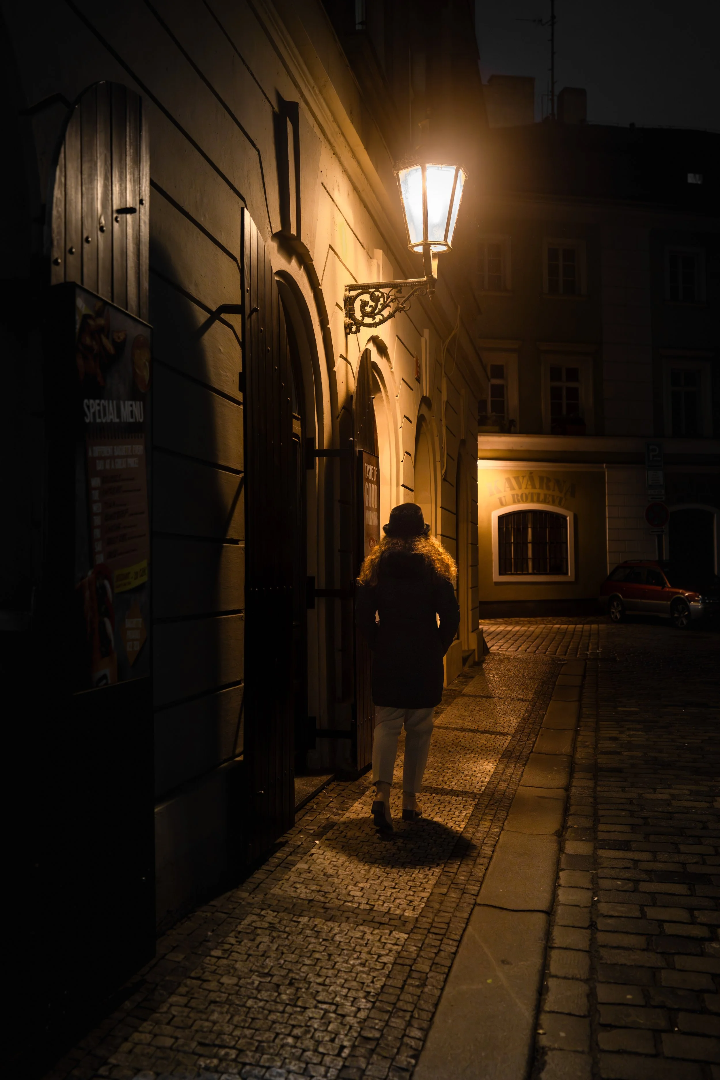 A woman walking on a cobblestone sidewalk at night, illuminated by a street lamp, with historic buildings and a restaurant sign in the background.