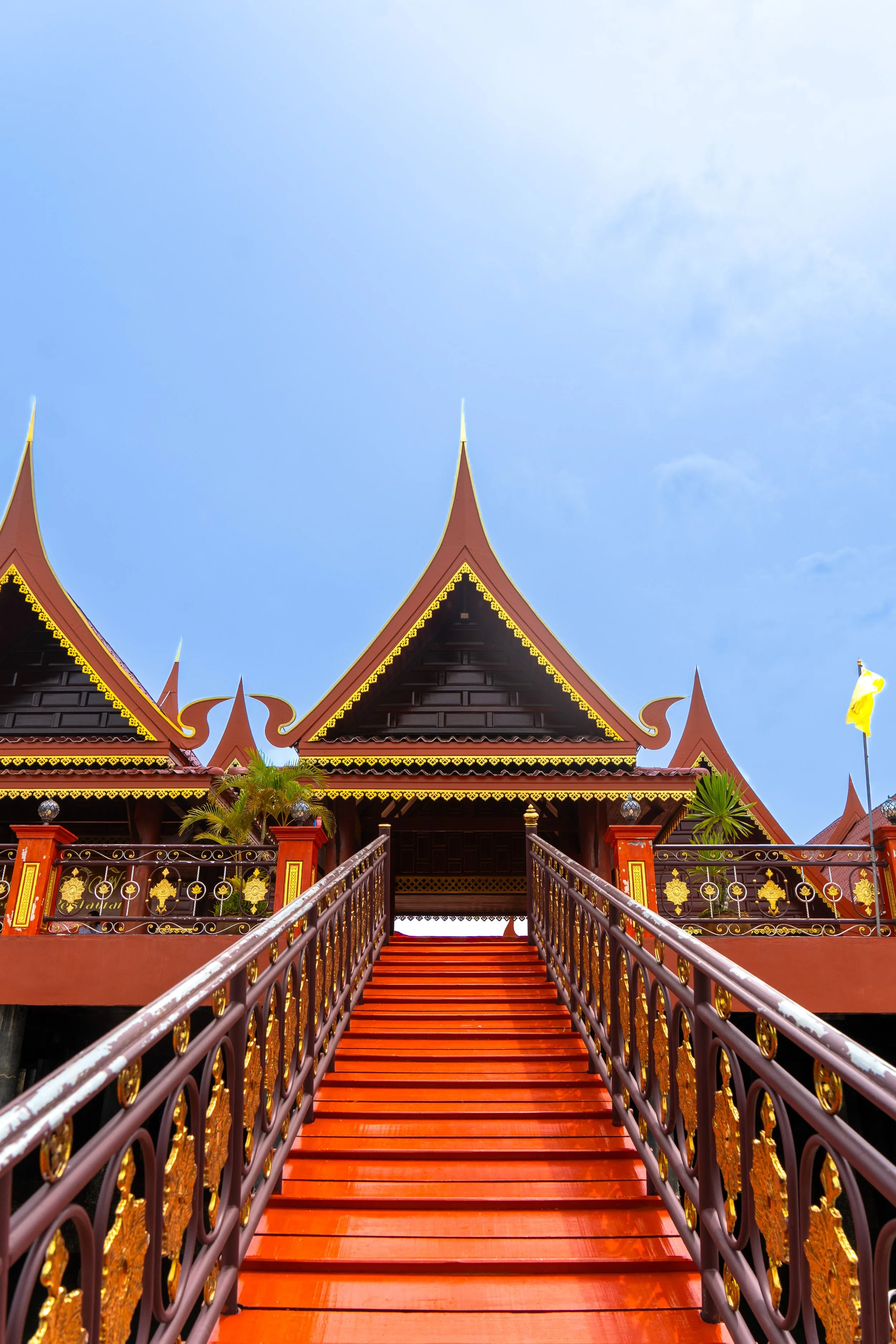 A traditional Thai temple with ornate red, gold, and dark wood architecture, a staircase leading up to the entrance, and a clear blue sky overhead.