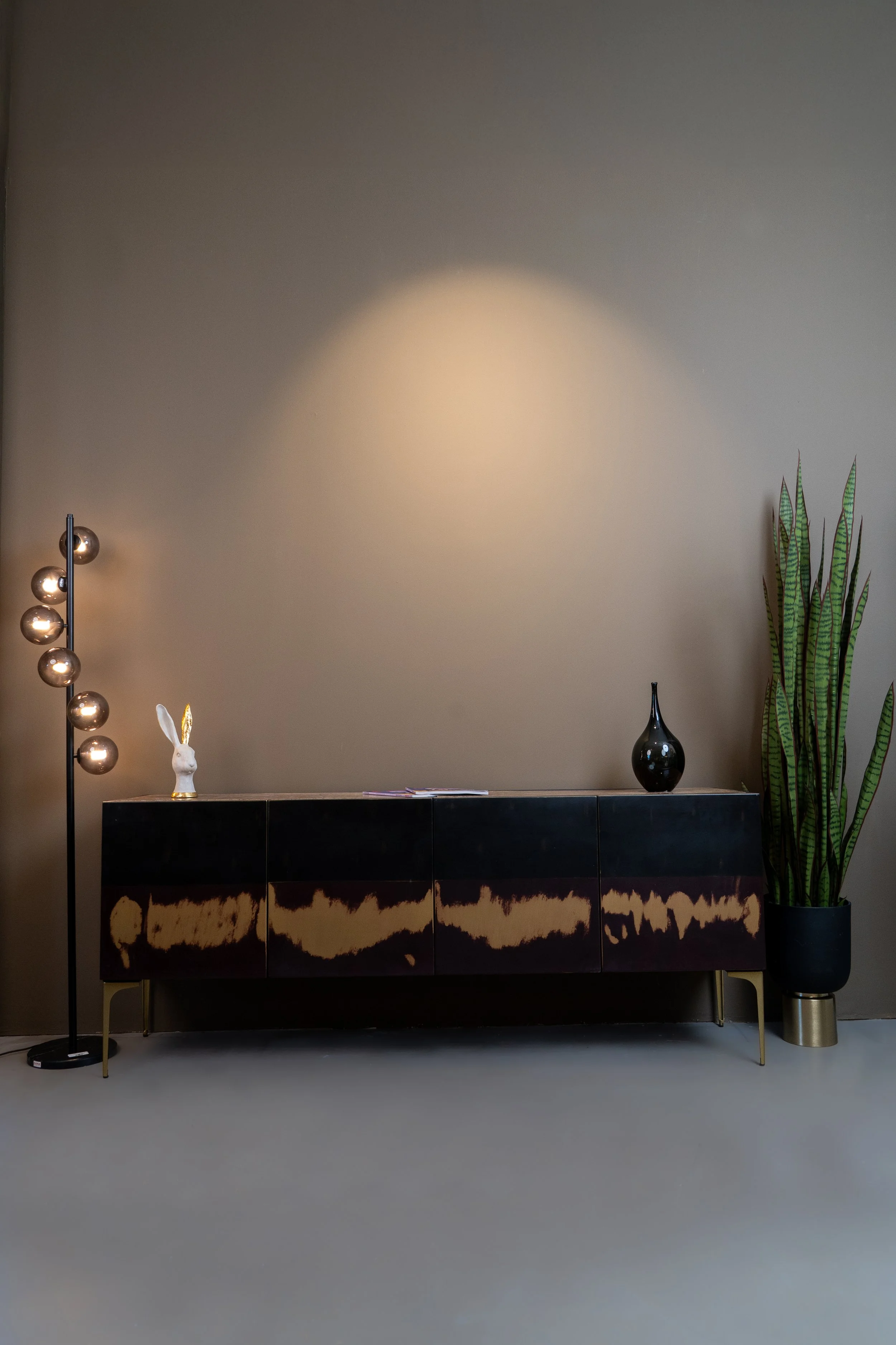 Minimalist living room with a black sideboard, a tall lamp with multiple bulbs, a decorative bunny head, a black vase, and a large potted snake plant, against a plain brown wall.