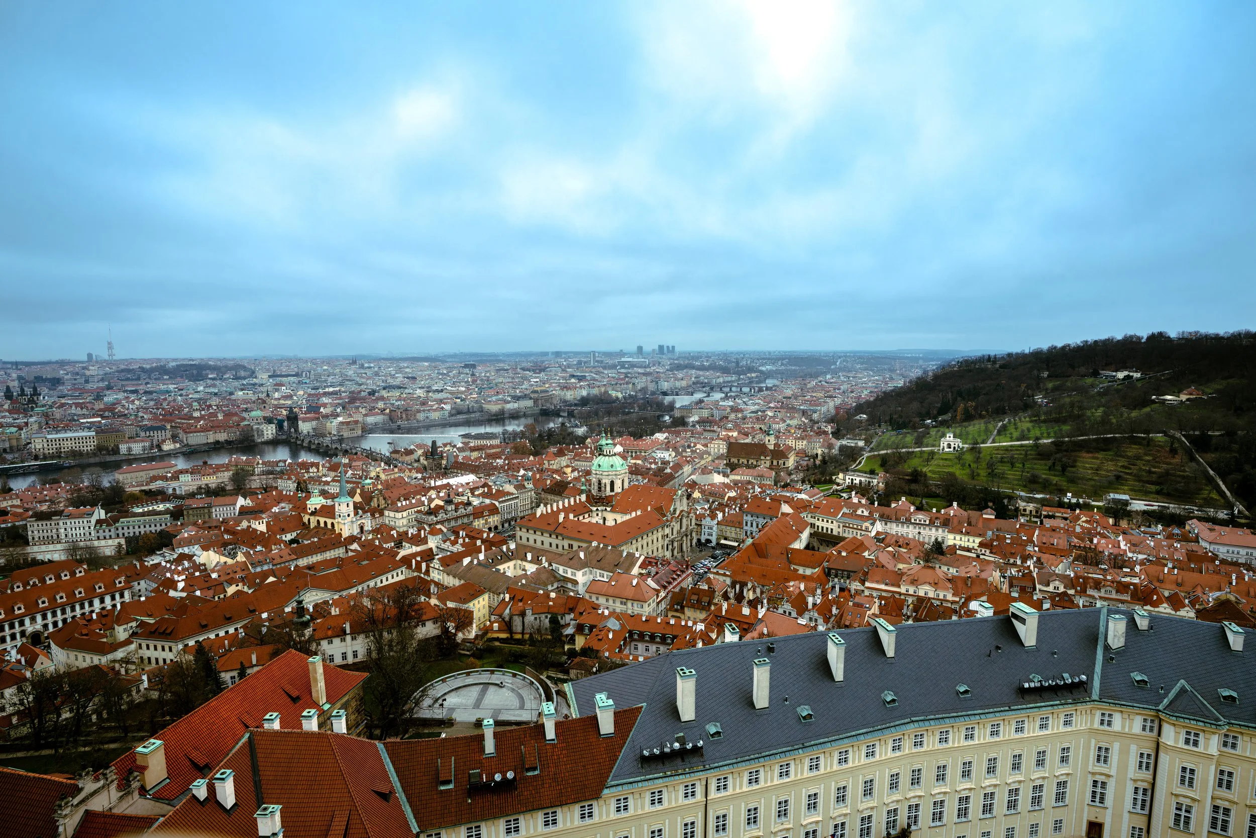 Aerial view of a European city with red-tiled roofs, a river running through the city, and a hill with greenery on the right side.
