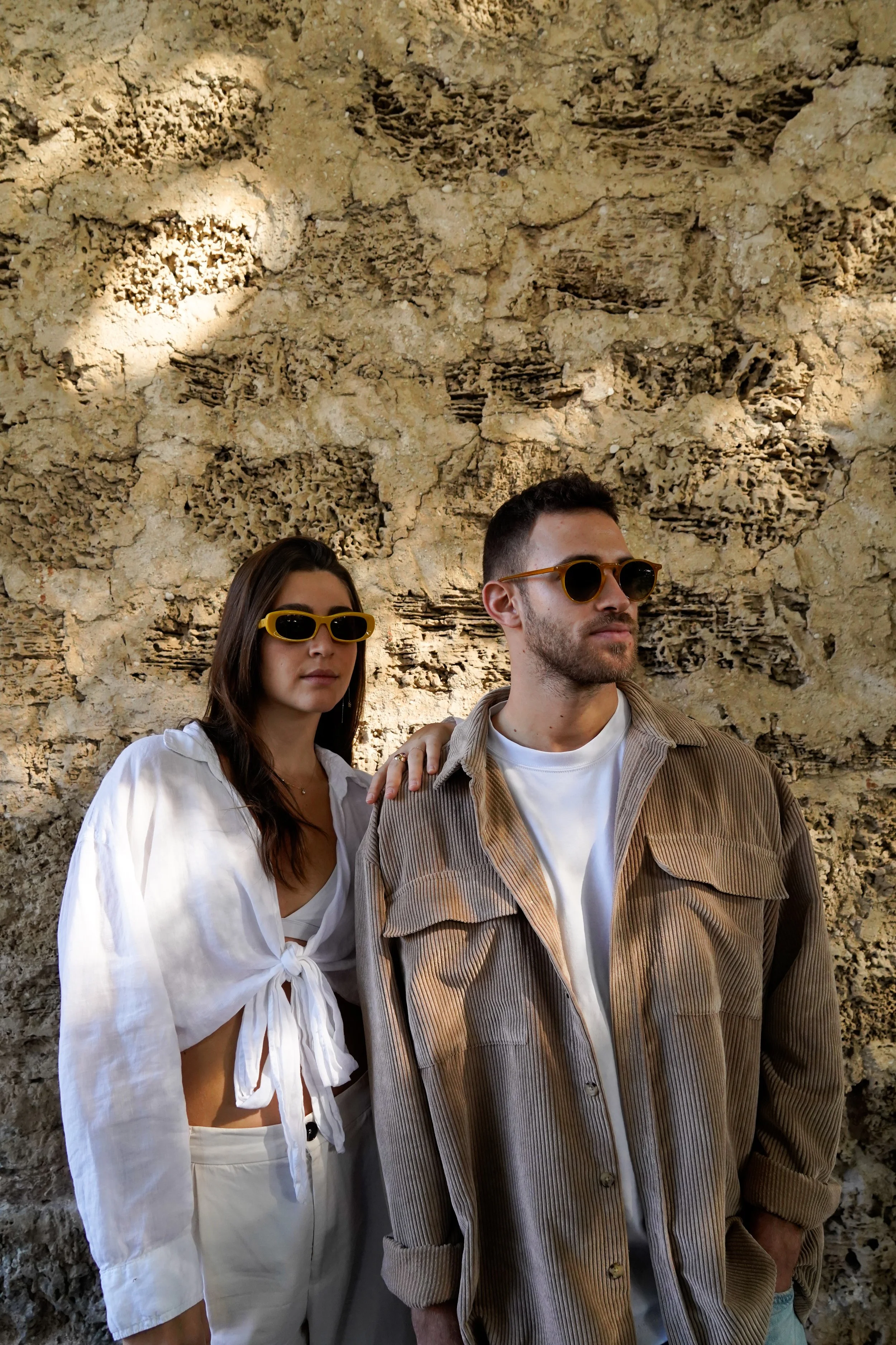 A man and woman standing in front of a textured beige stone wall, both wearing sunglasses, with the woman resting her hand on the man's shoulder.