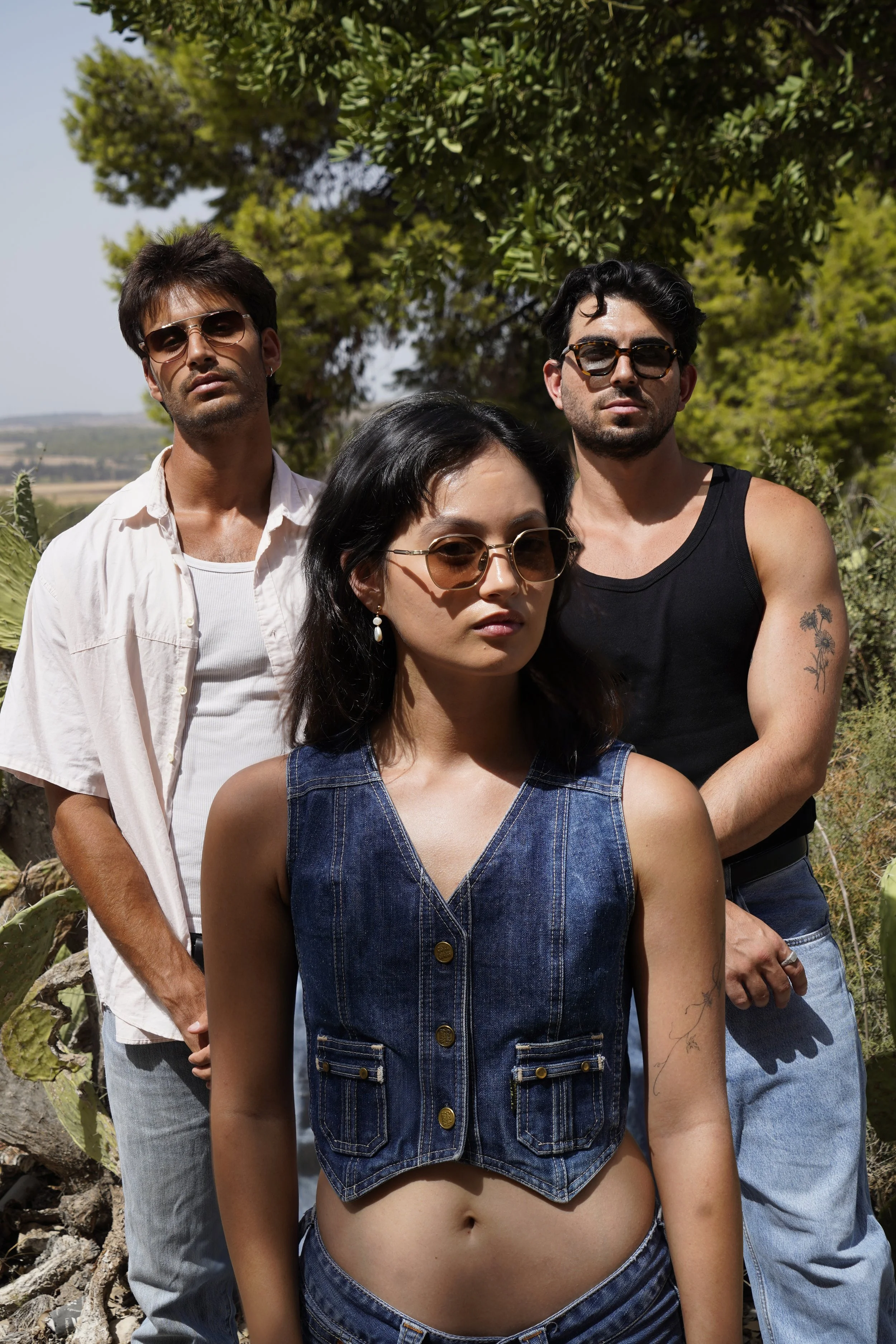 Three young adults outdoors: a woman in front wearing sunglasses and a sleeveless denim vest, two men behind her wearing sunglasses, one in a black tank top and the other in a white shirt, with trees and clear sky in the background.