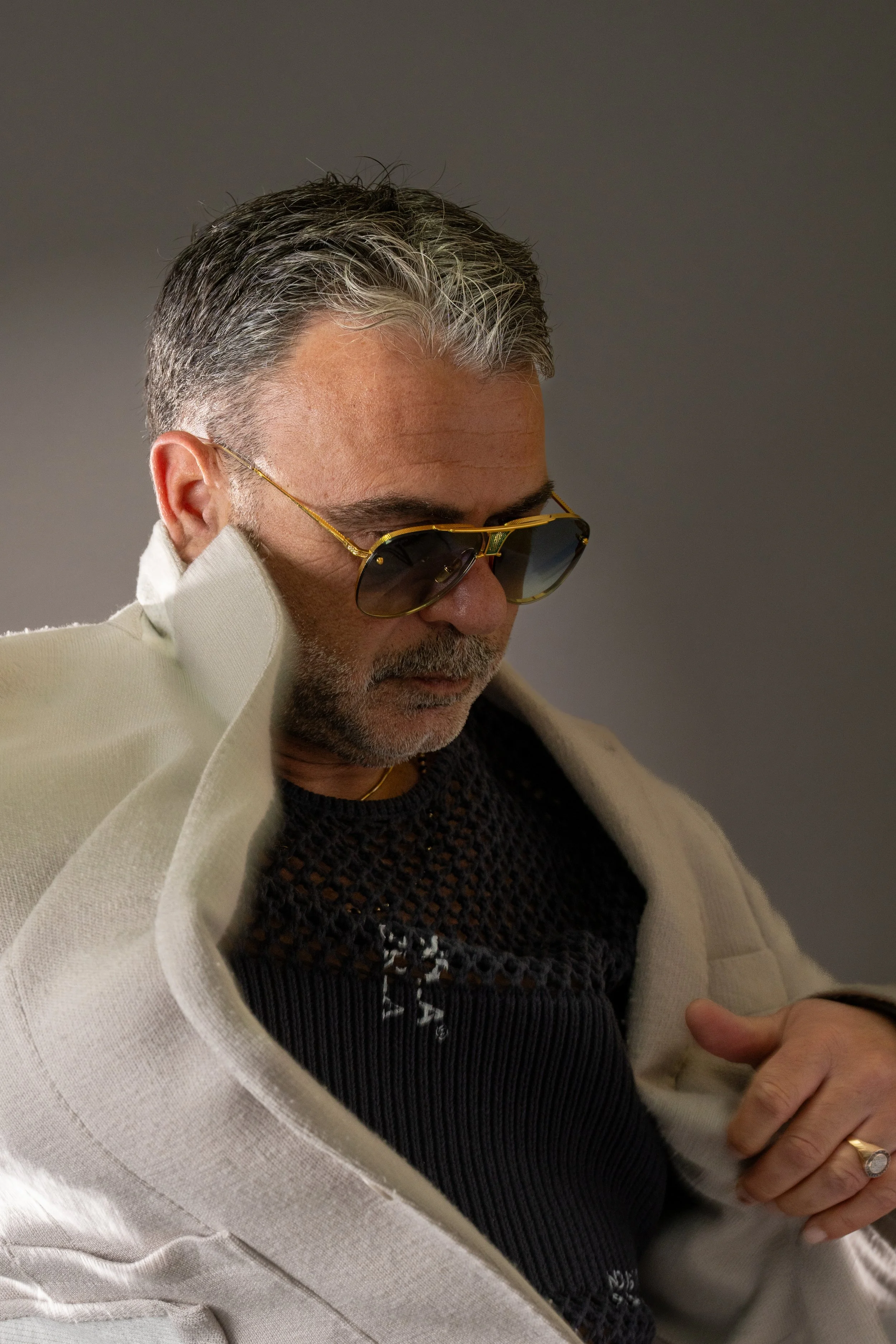A middle-aged man with gray hair and beard wearing dark sunglasses, a black knit sweater, and a light-colored jacket, looking down with a serious expression.
