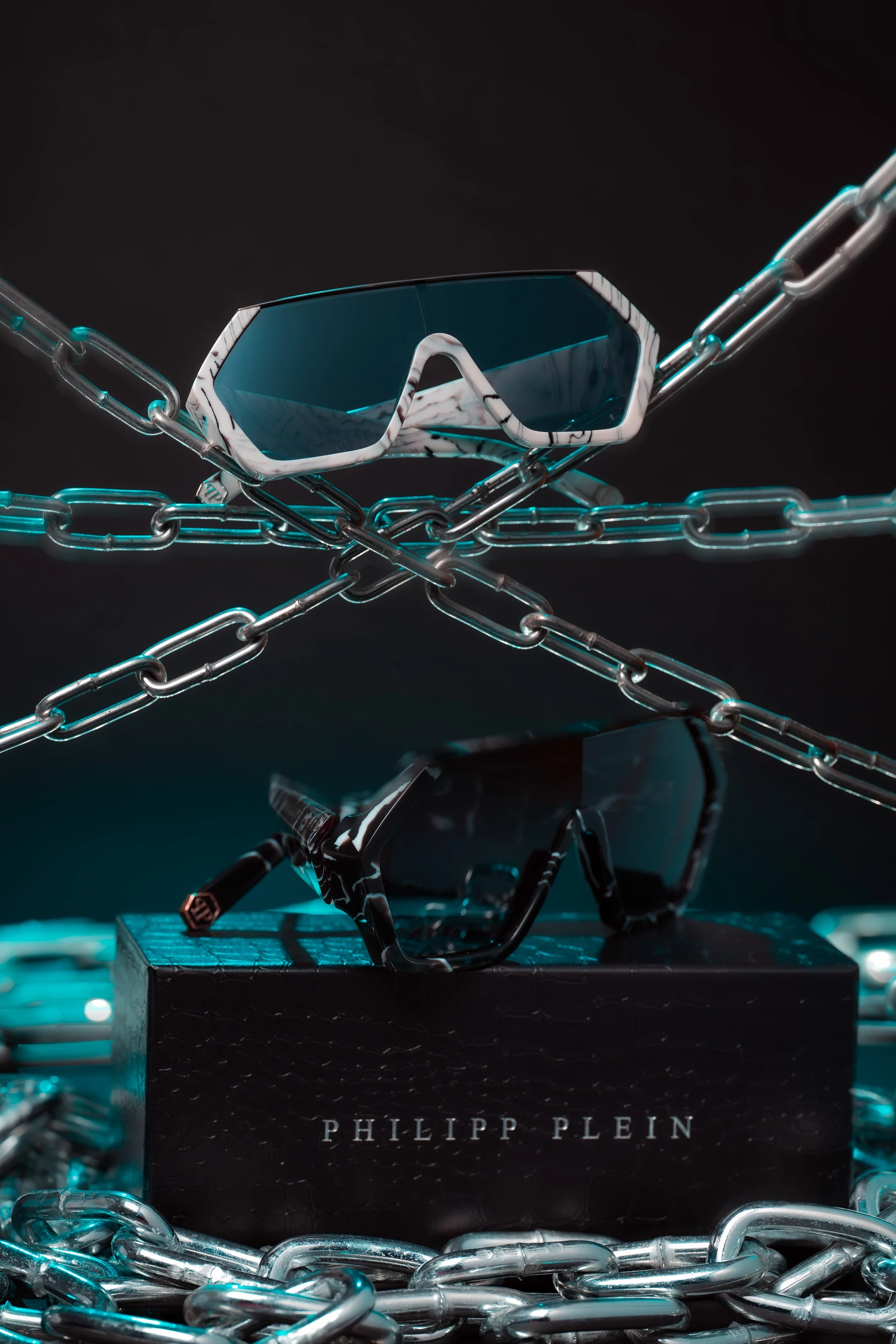 Pair of Philipp Plein sunglasses on top of a black box with the brand's name, surrounded by silver chains and another pair of sunglasses, all against a dark background.
