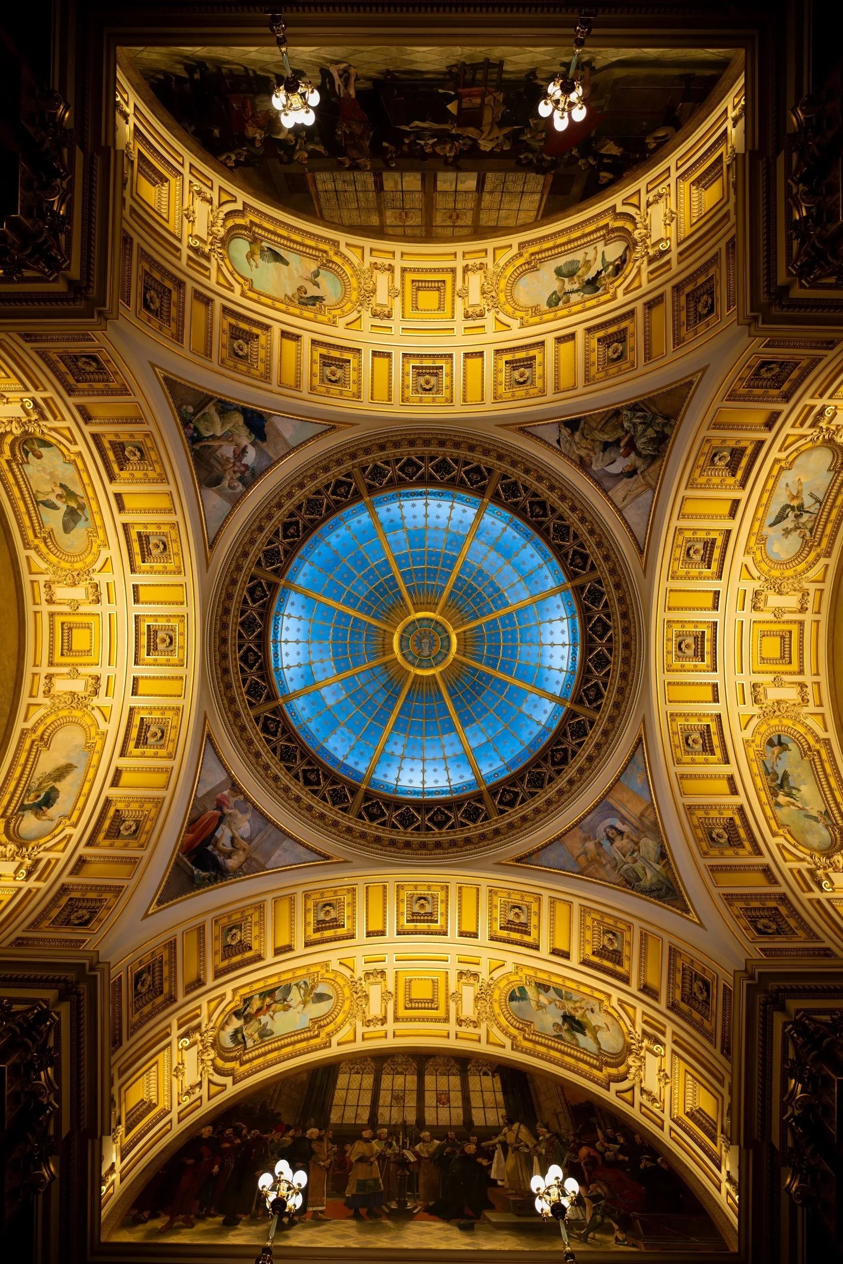 Interior view of a domed ceiling with a blue stained glass center, surrounded by ornate gilded decorations, paintings, sculptures, and chandeliers.