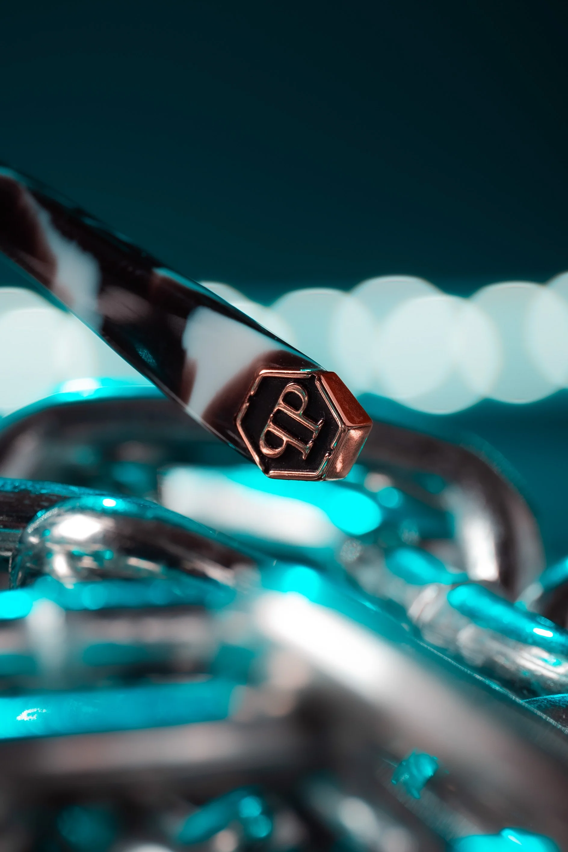 Close-up of a metallic pen with an engraved logo, resting on a pile of interconnected silver chains, with a bokeh background of soft white lights.