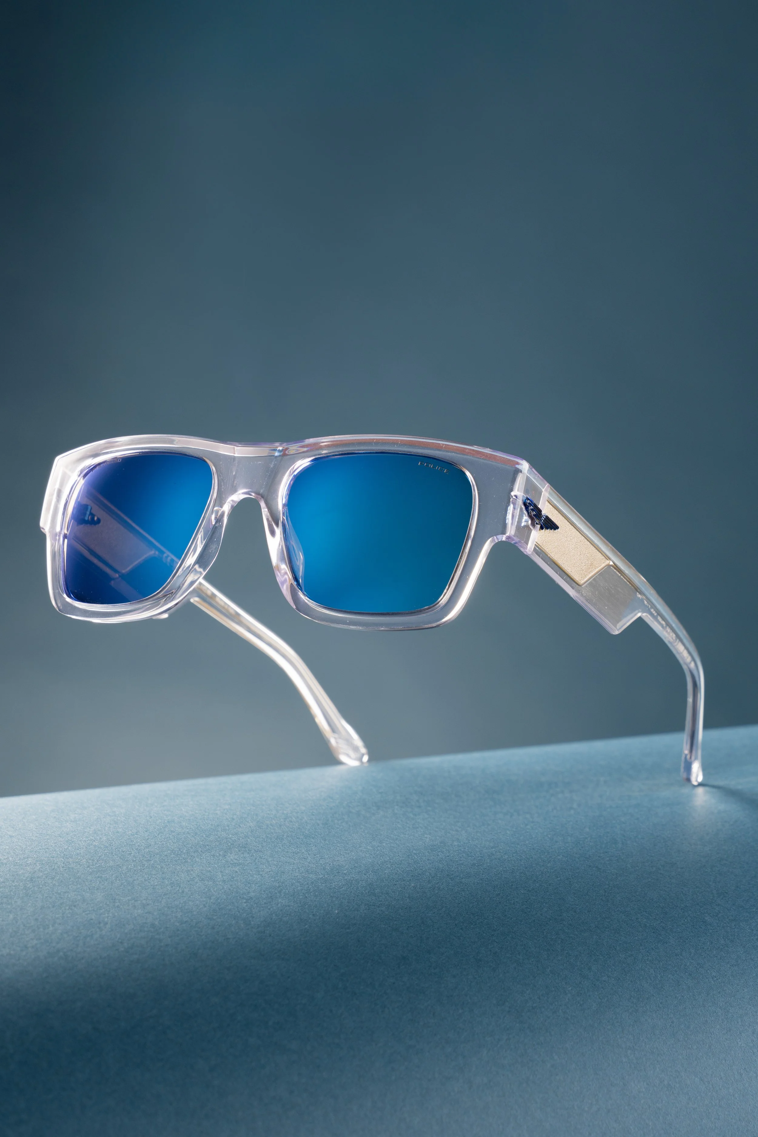 Clear pair of sunglasses with blue lenses placed on a teal surface with a background that transitions from teal to dark blue.