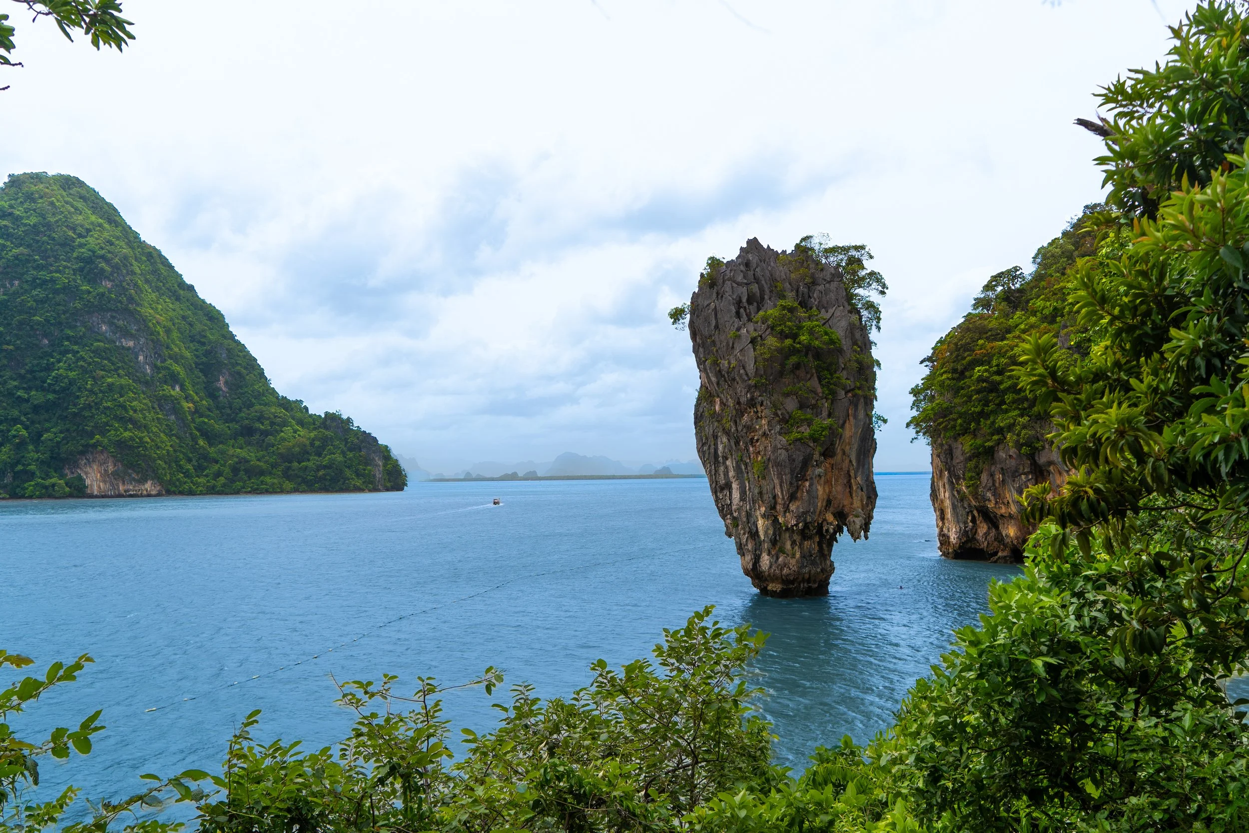 Sea with limestone islands, rocky formations, lush green foliage, and a boat in the distance.