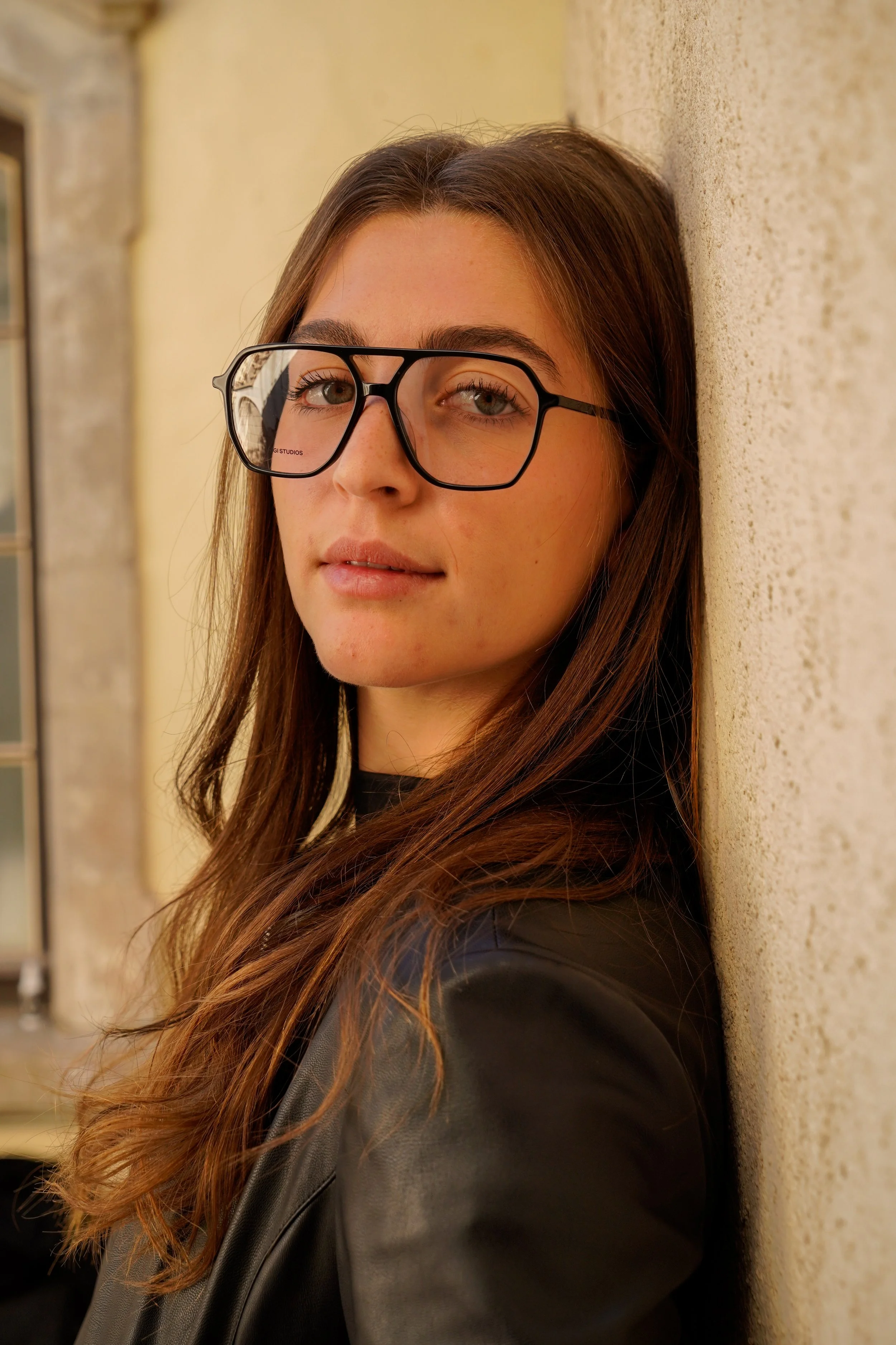 A young woman with brown hair and glasses leaning against a beige brick wall outdoors.