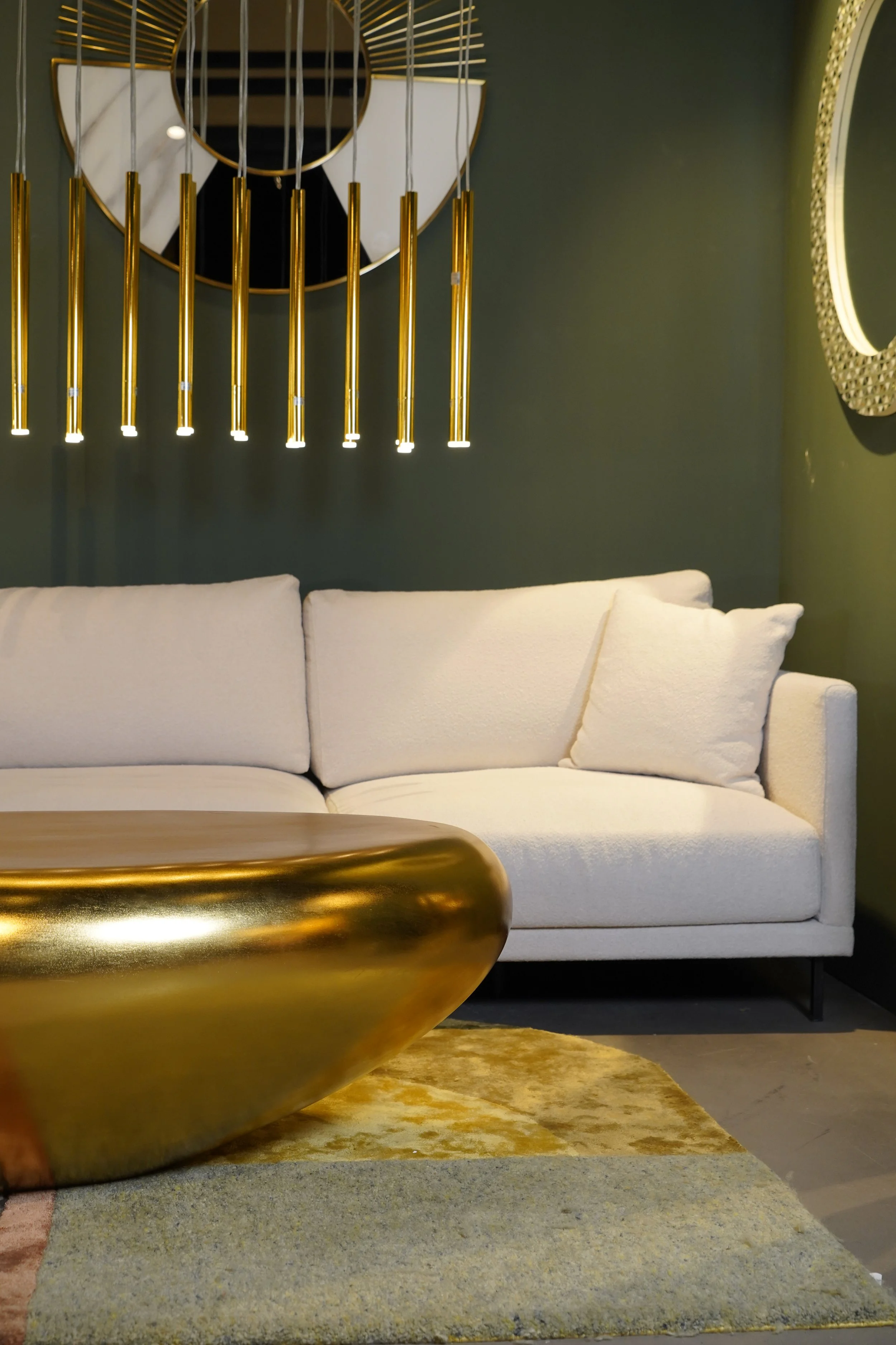 A modern living room with a white sofa, a gold metallic coffee table, a dark green wall, a circular gold wall mirror, and a contemporary gold pendant chandelier.