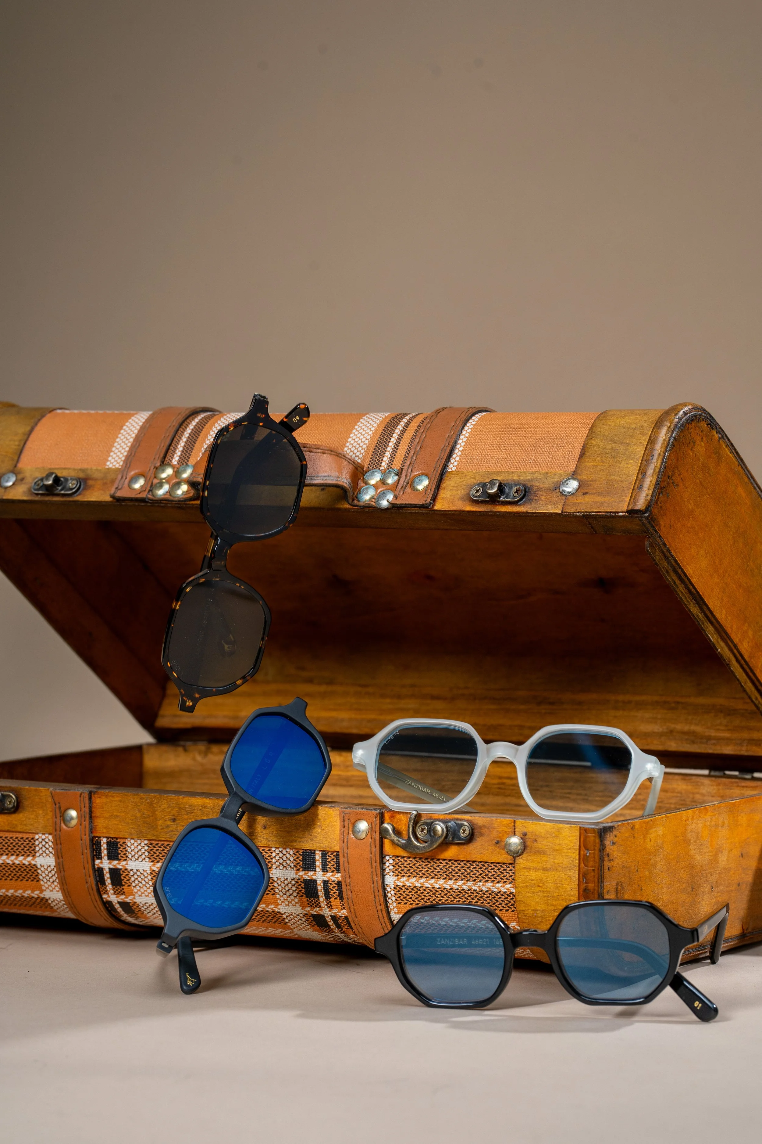 A collection of sunglasses in black, blue, and clear styles displayed in and around an open vintage wooden suitcase with decorative fabric and metal accents.