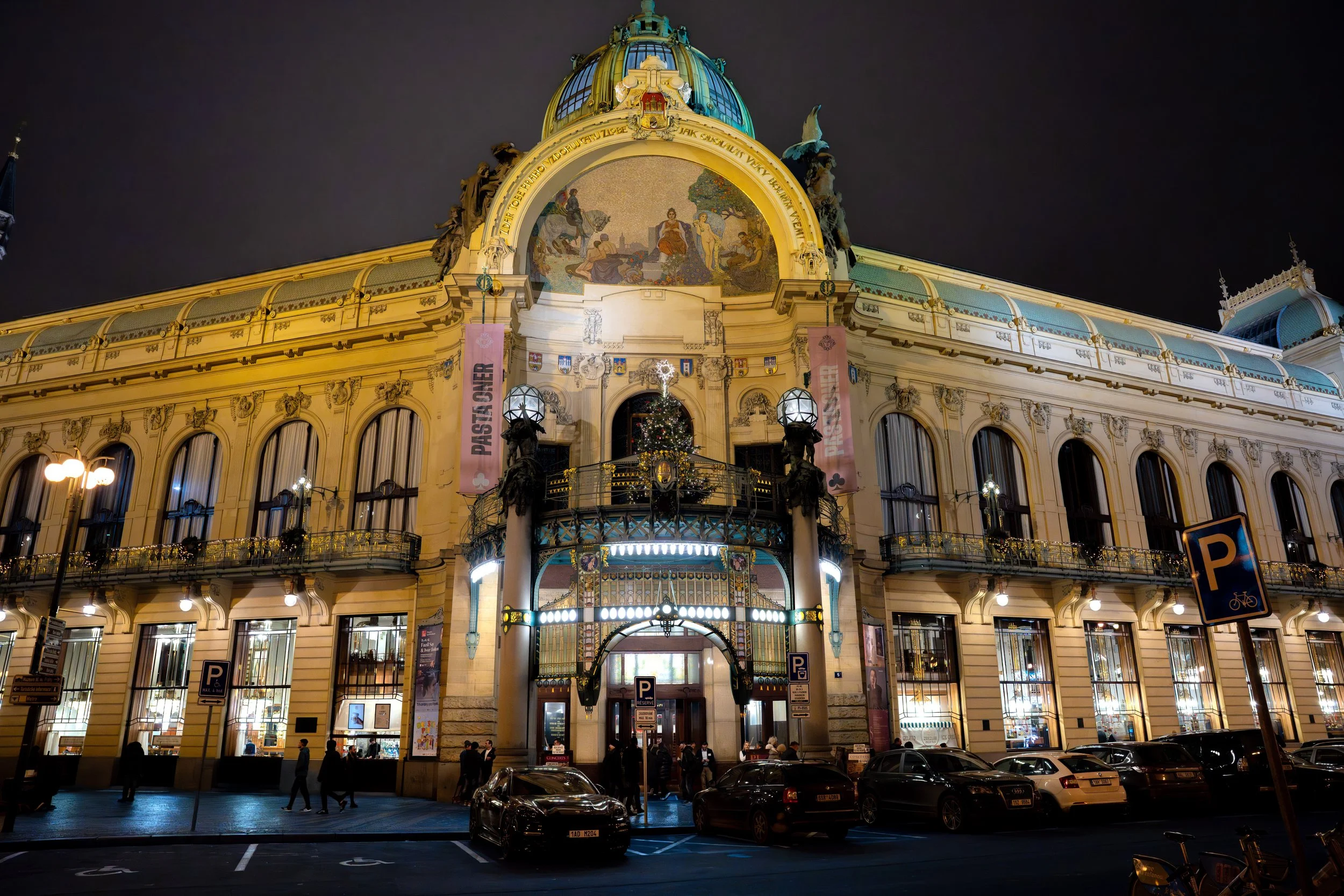 Night view of a historic, ornate building decorated with a Christmas tree and festive lights, with parked cars and pedestrians in front.