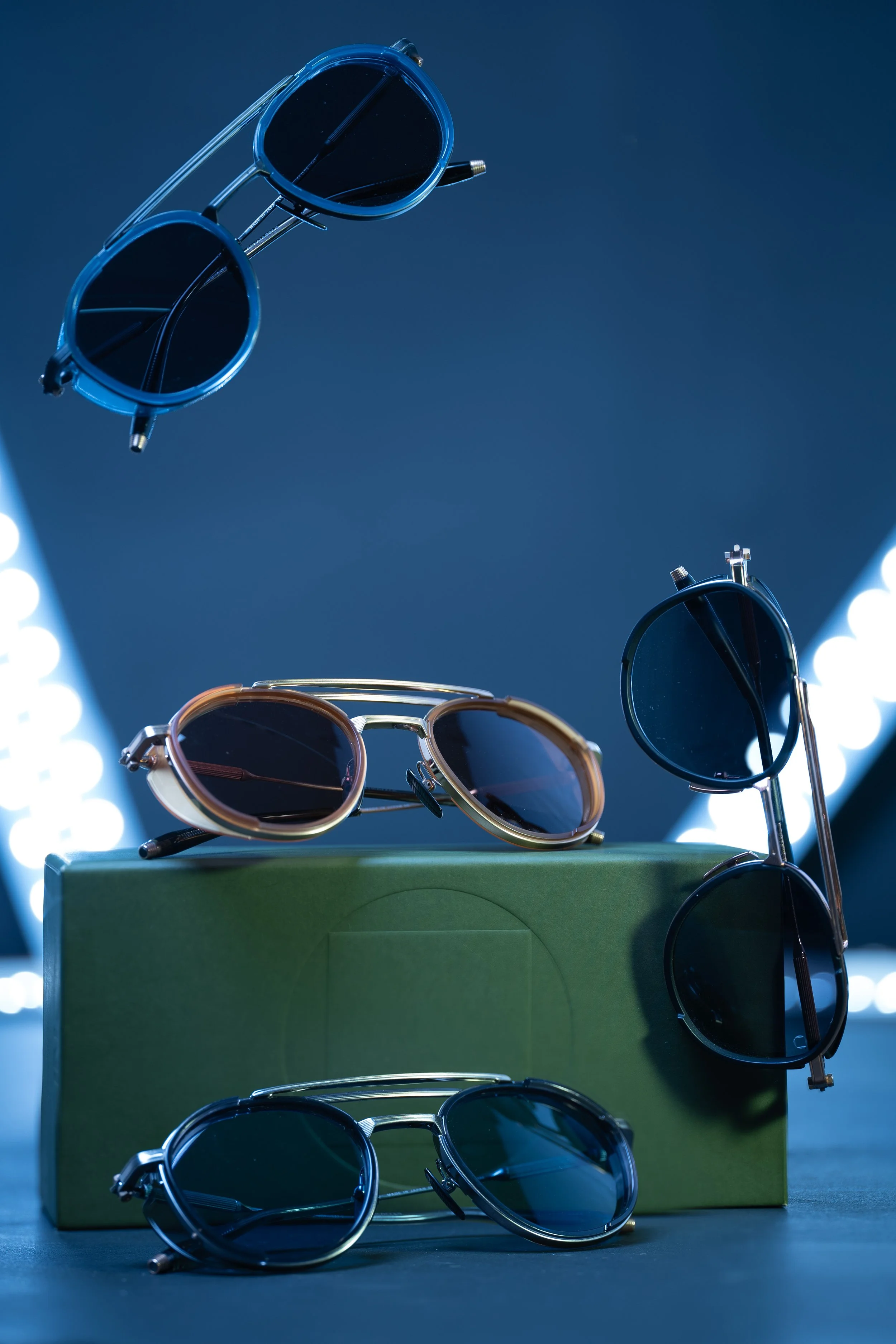 Multiple pairs of sunglasses placed on and around a green box against a dark background with bright lights.