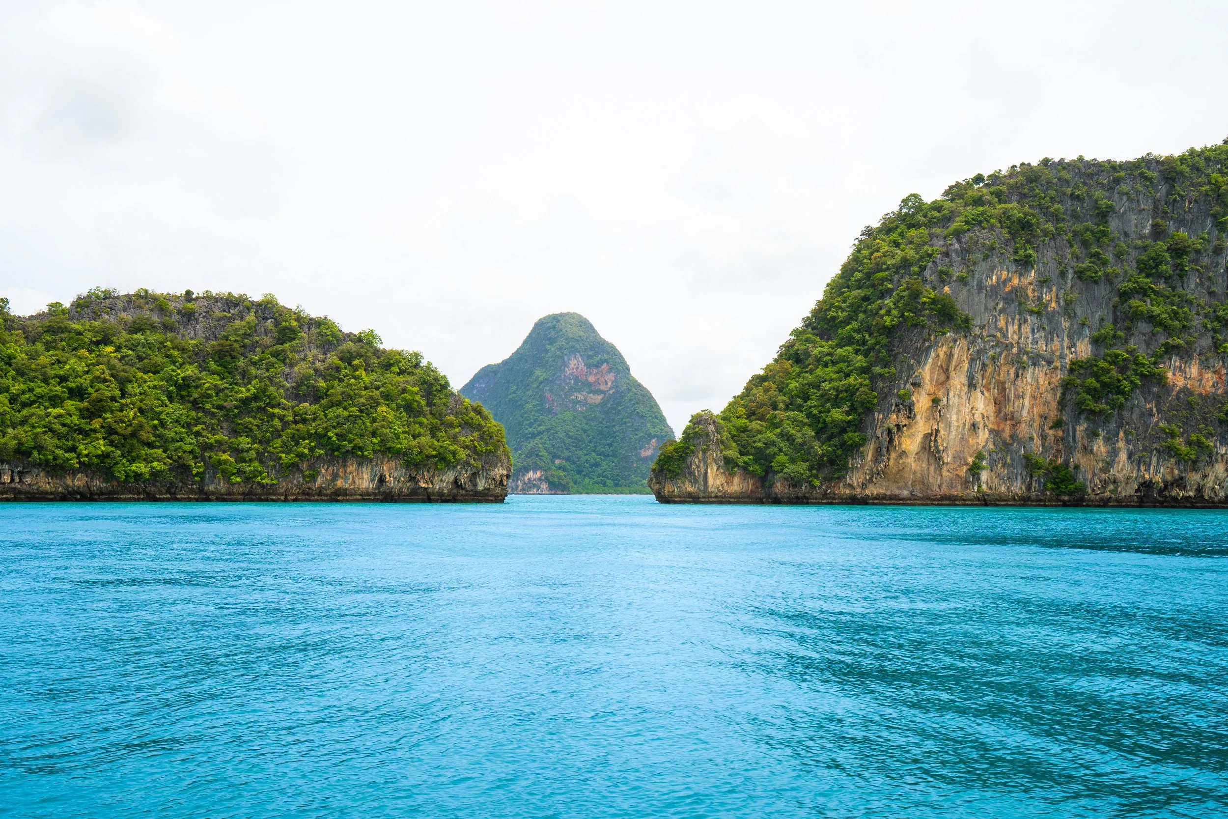 Tropical seascape with turquoise water surrounded by lush green islands and limestone cliffs, and a mountain in the distance under a cloudy sky.
