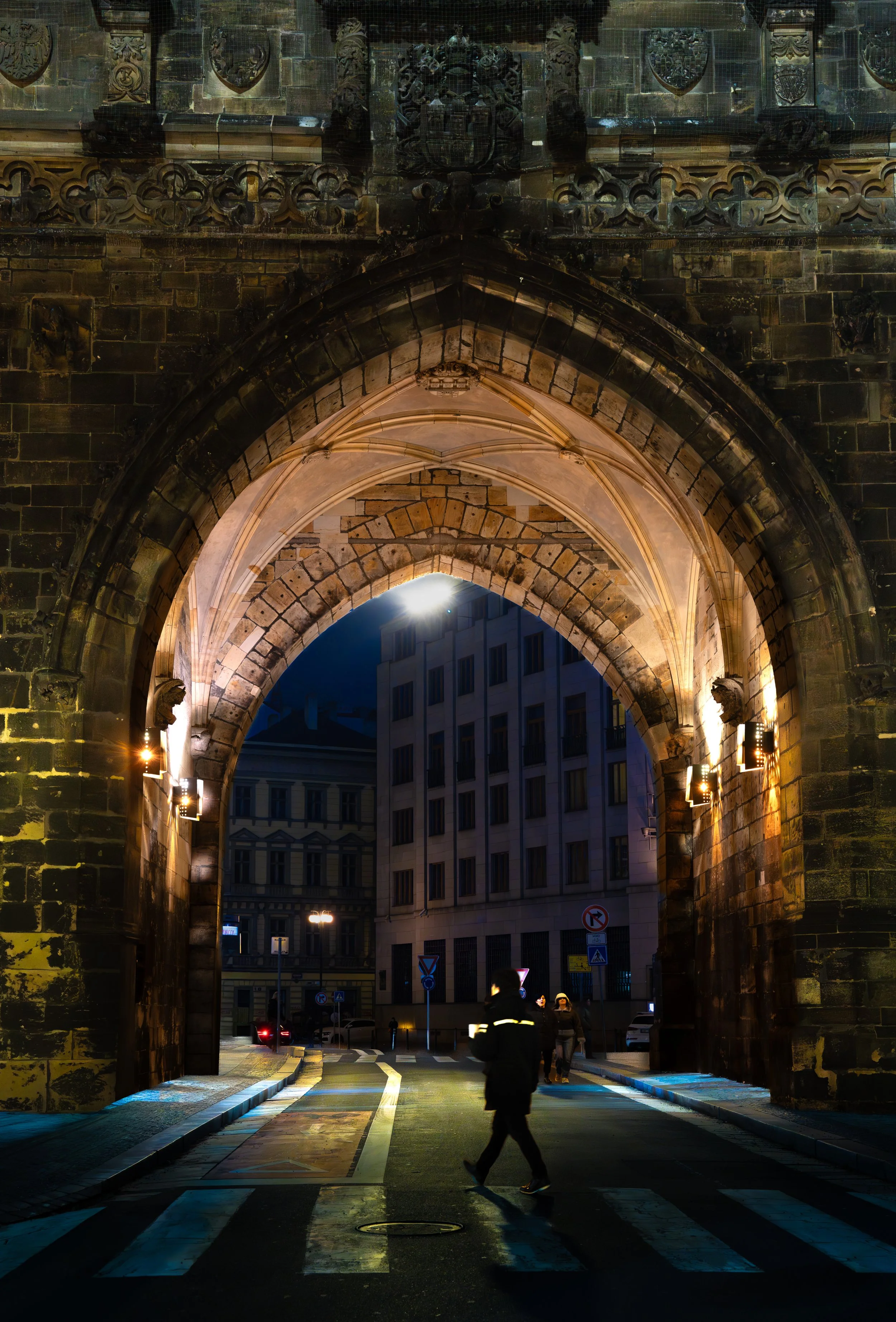 Night scene of a historic stone archway with gothic architectural details, illuminated by warm lights, leading into a city street with pedestrians.