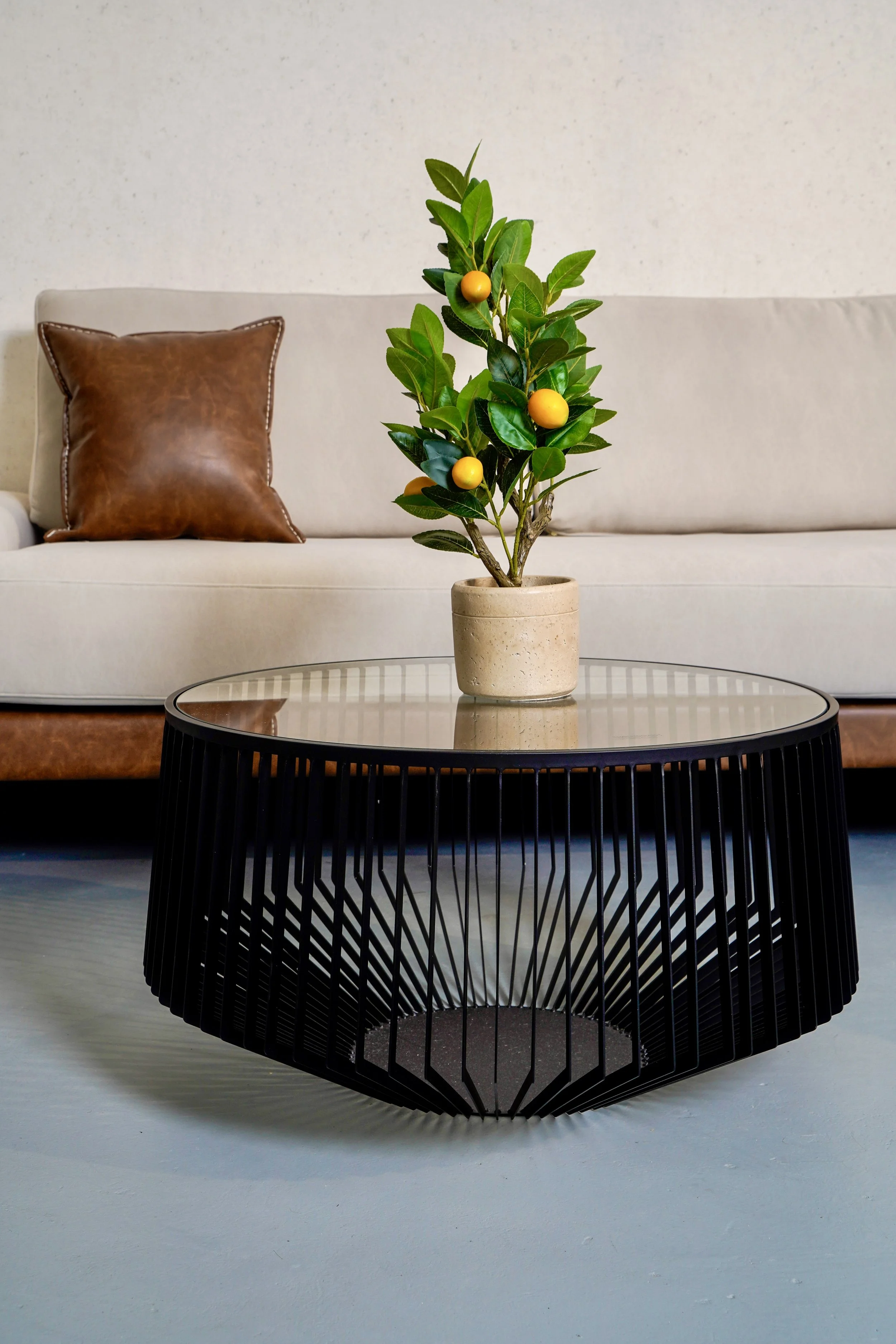 Modern living room with a round glass-top coffee table and a potted lemon tree, beige sofa, and a brown leather pillow.
