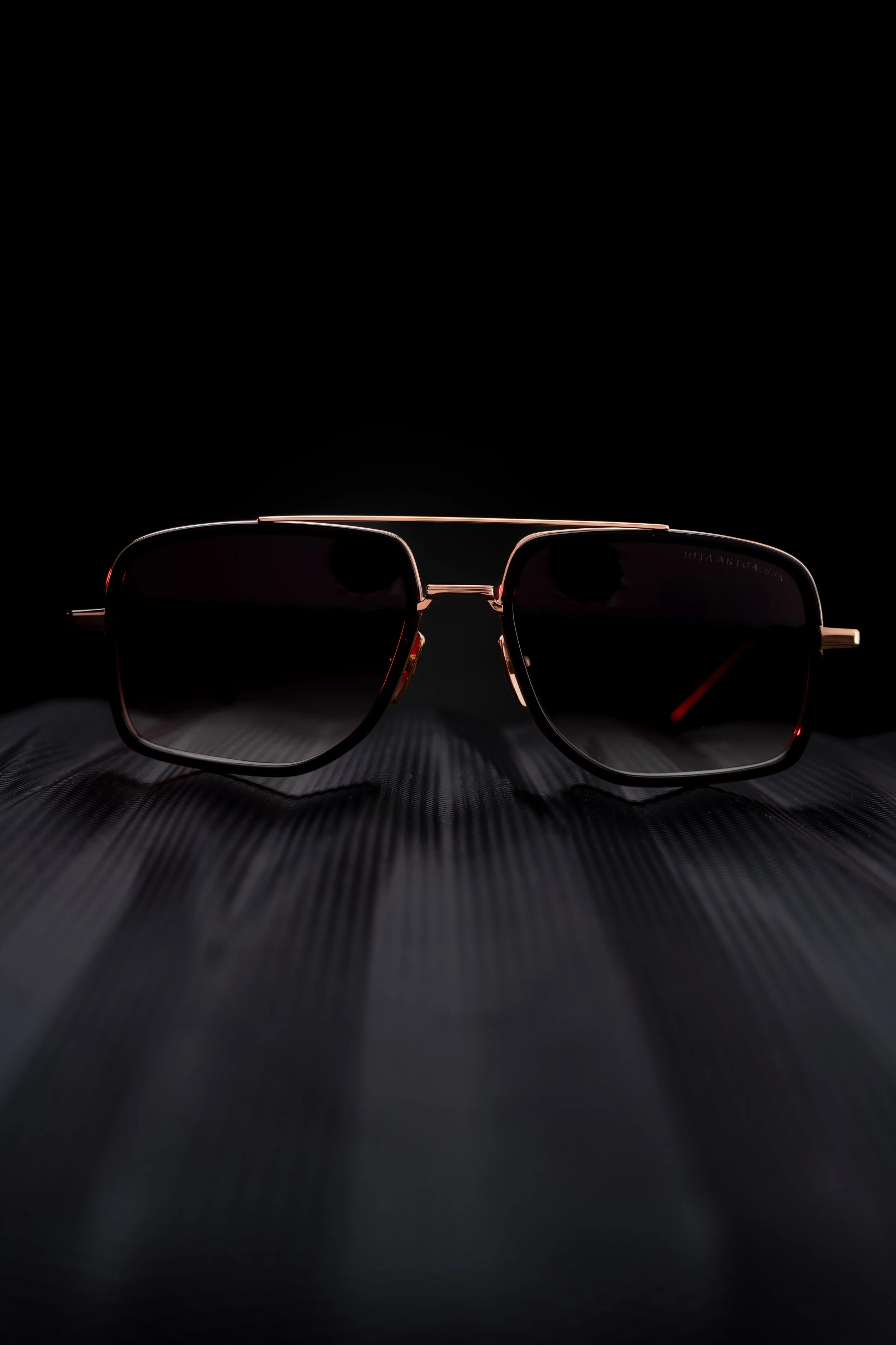 Pair of sunglasses with dark lenses and a gold frame resting on a textured black surface against a dark background.