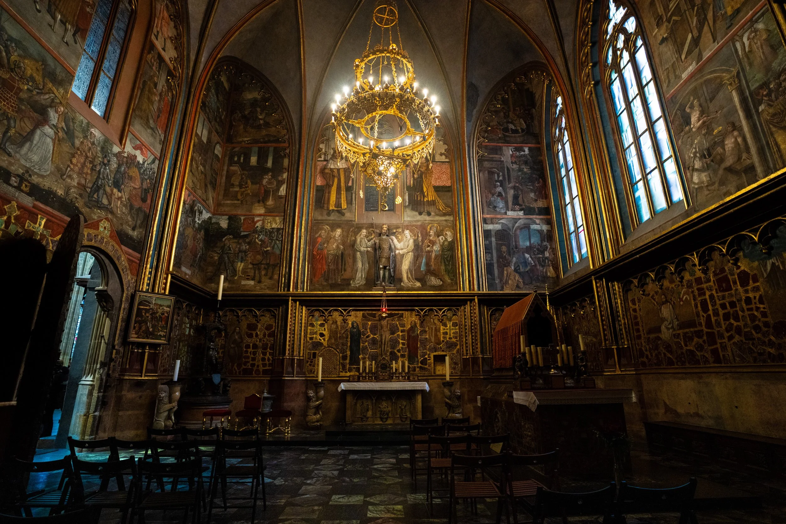 Interior of a church or chapel with ornate gold accents, stained glass windows, a chandelier, religious murals, an altar, and chairs for worshippers.