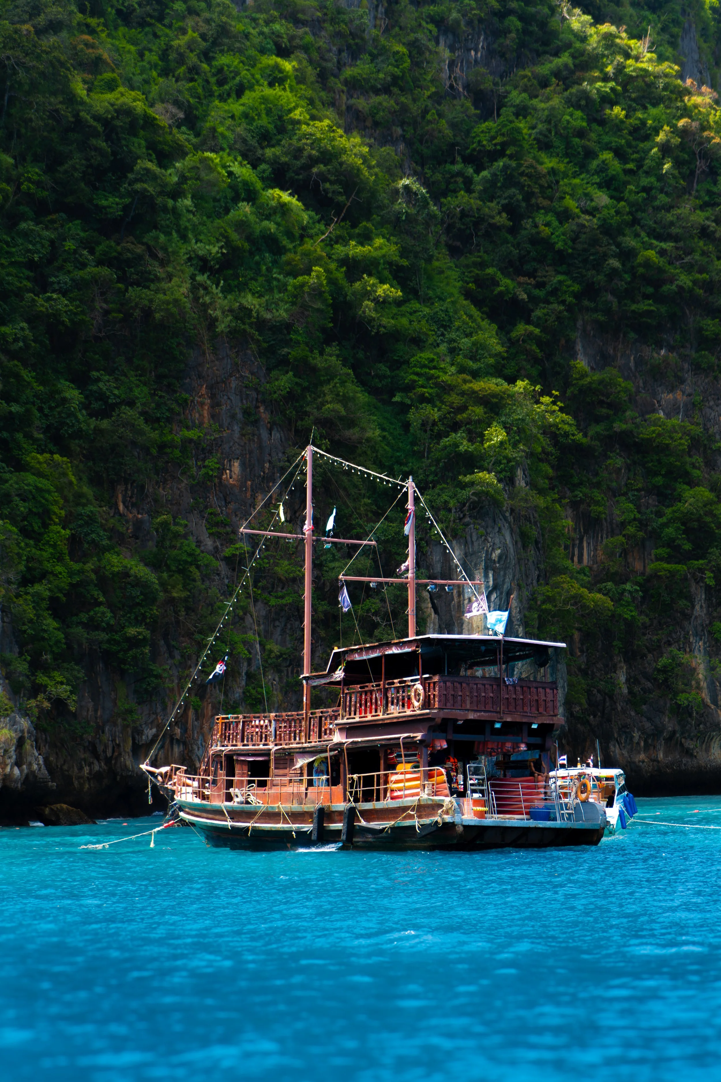 A wooden boat with two masts floating on clear blue water near a lush green cliff.