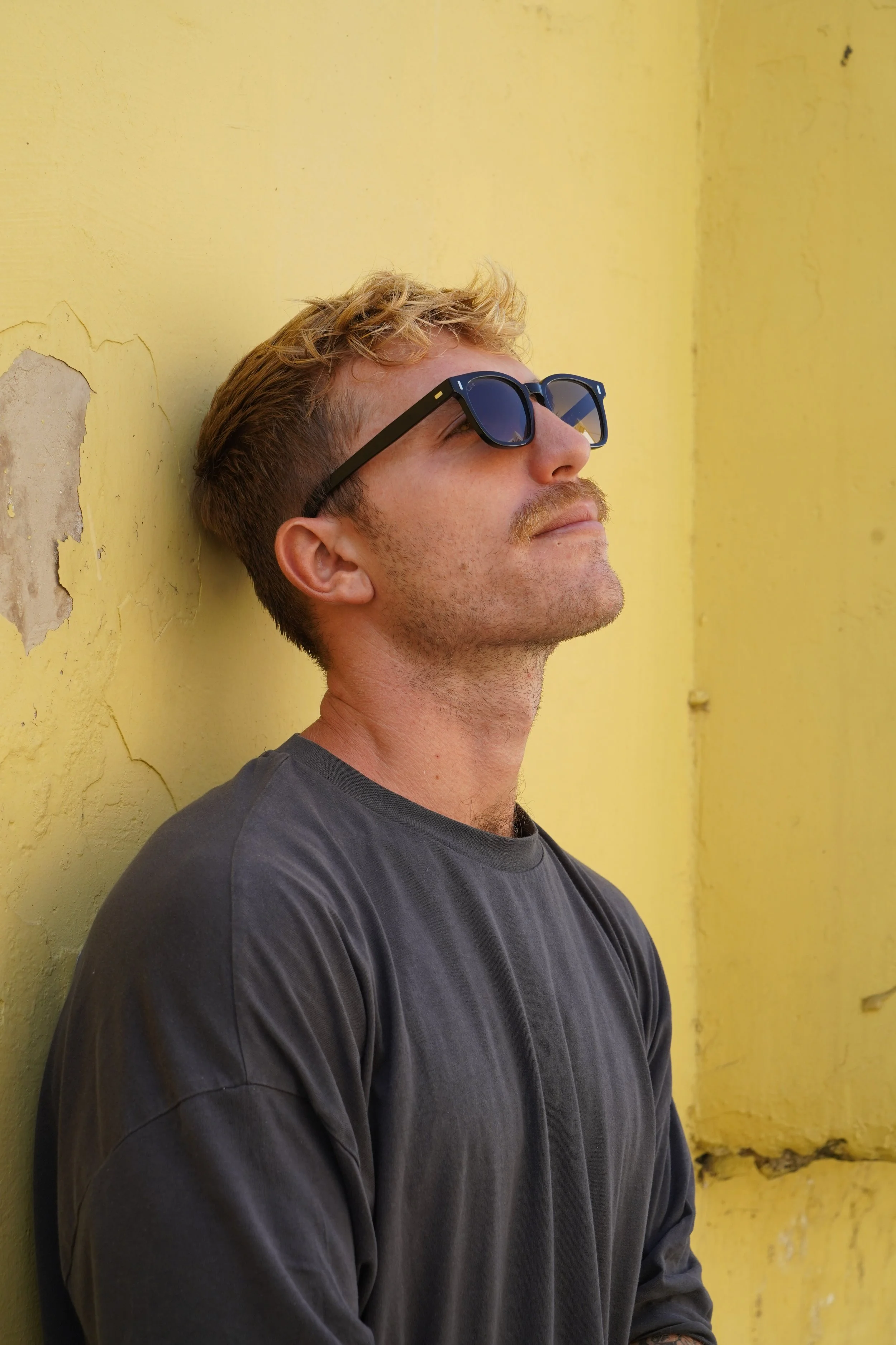 A man with blond hair and sunglasses leaning against a yellow wall with peeling paint.