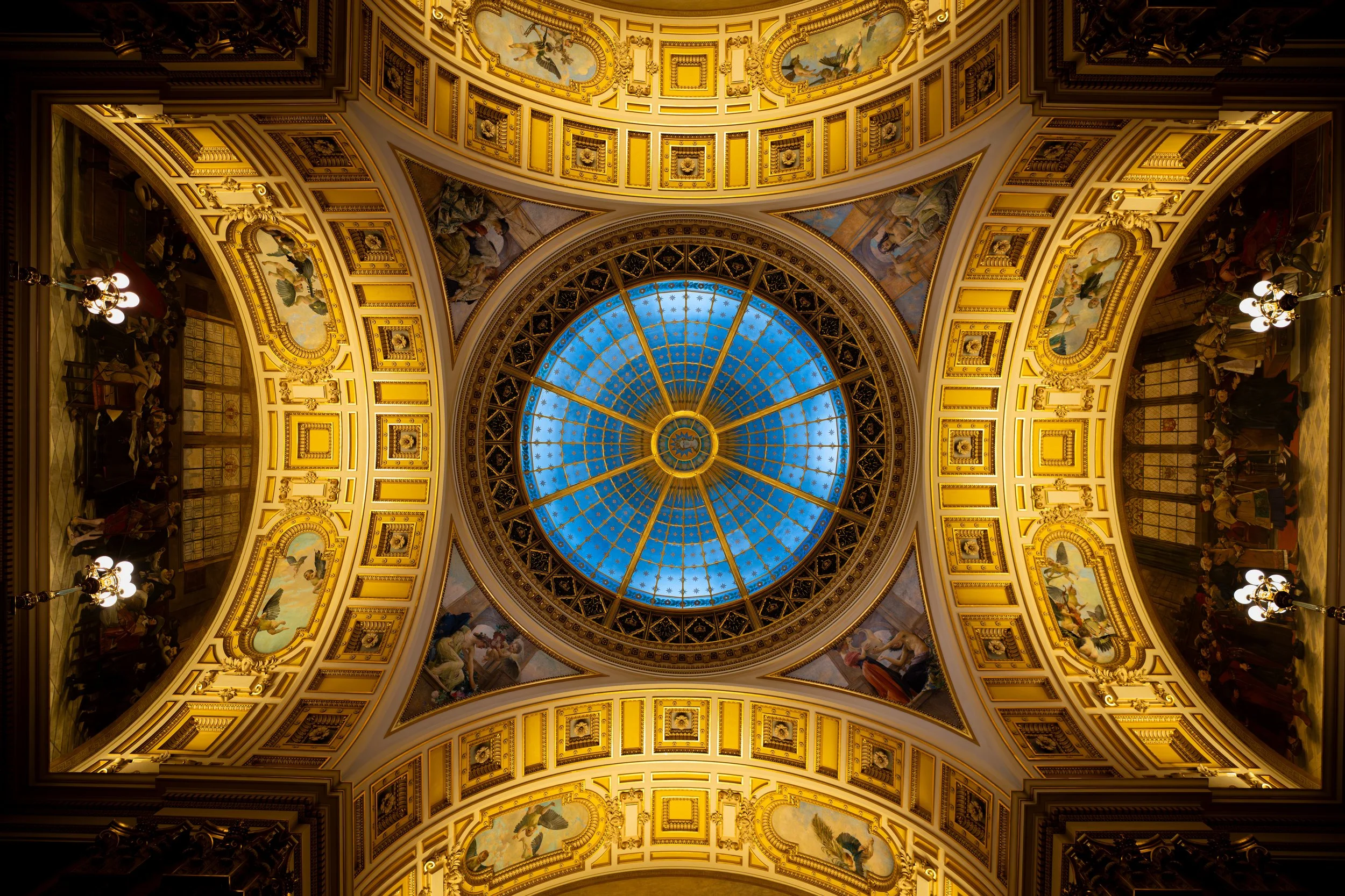Interior view of a grand, ornate ceiling with a central blue stained glass dome surrounded by gold detailing and painted murals.
